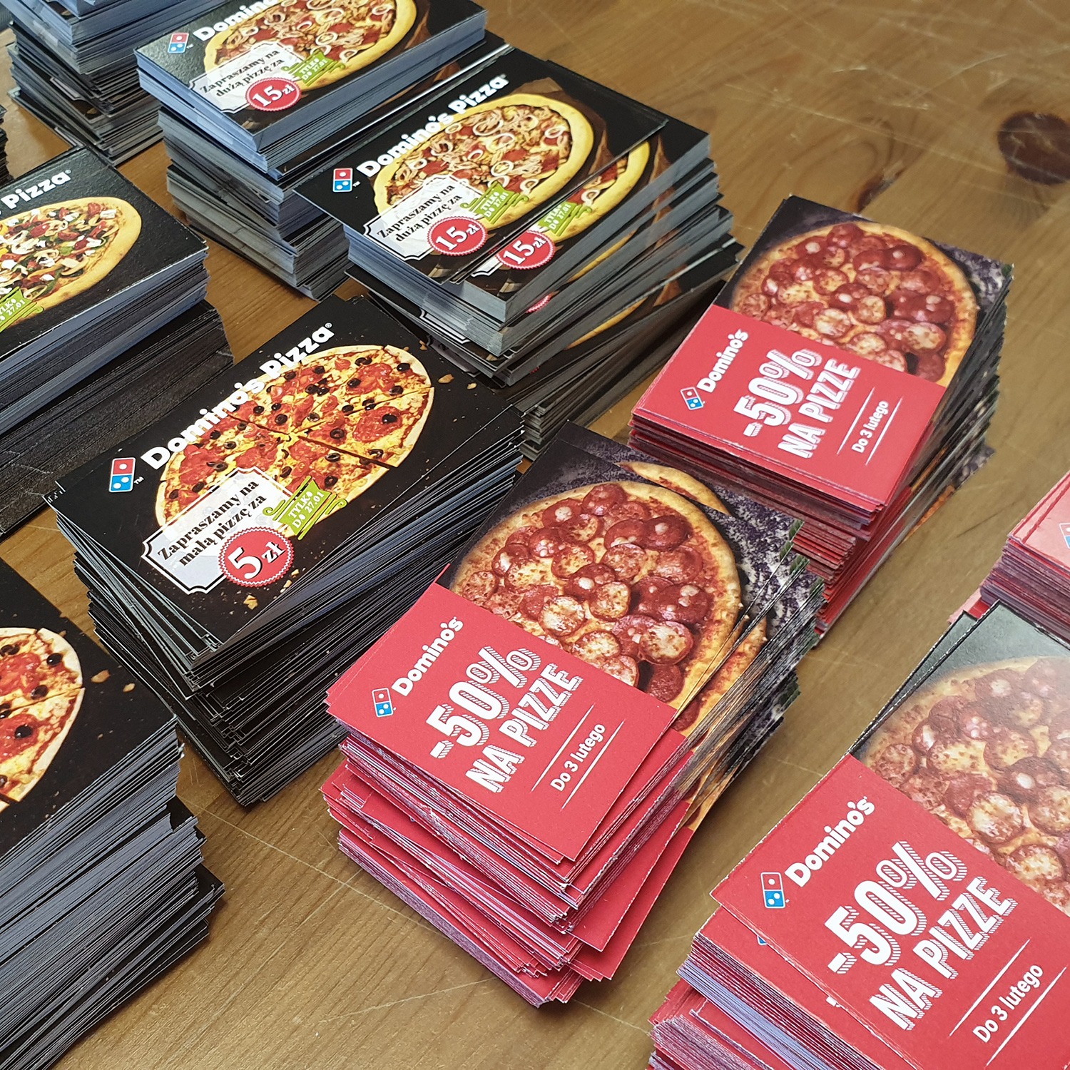 Stosy kartonowych materiałów reklamowych z grafiką pizzy i logo Domino's, ułożone na drewnianym stole.