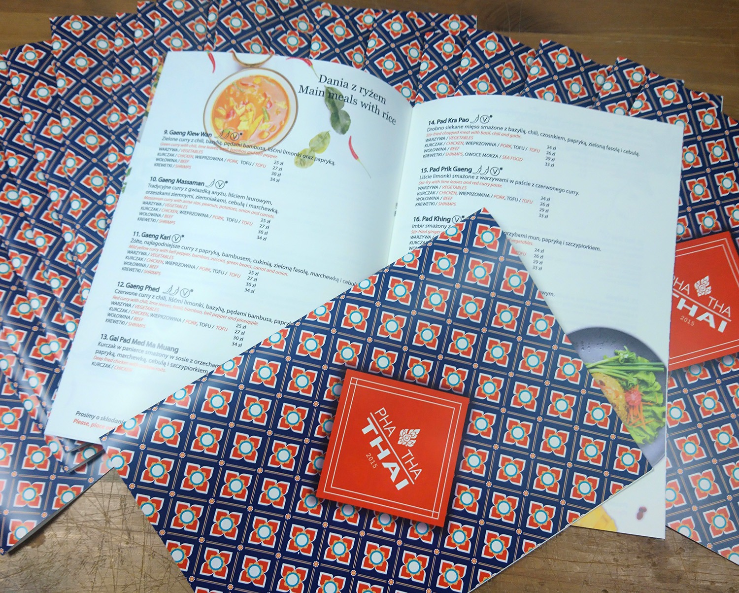 Rozłożone menu restauracji tajskiej z daniami i cenami, ozdobione motywem kwiatowym, obok leżą małe kwadratowe ulotki z logo restauracji.