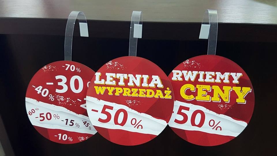 Trzy okrągłe woblery reklamowe z hasłami 'Letnia Wyprzedaż', 'Rwiemy Ceny' i informacjami o zniżkach, zawieszone na przezroczystych haczykach na tle ciemnej półki.