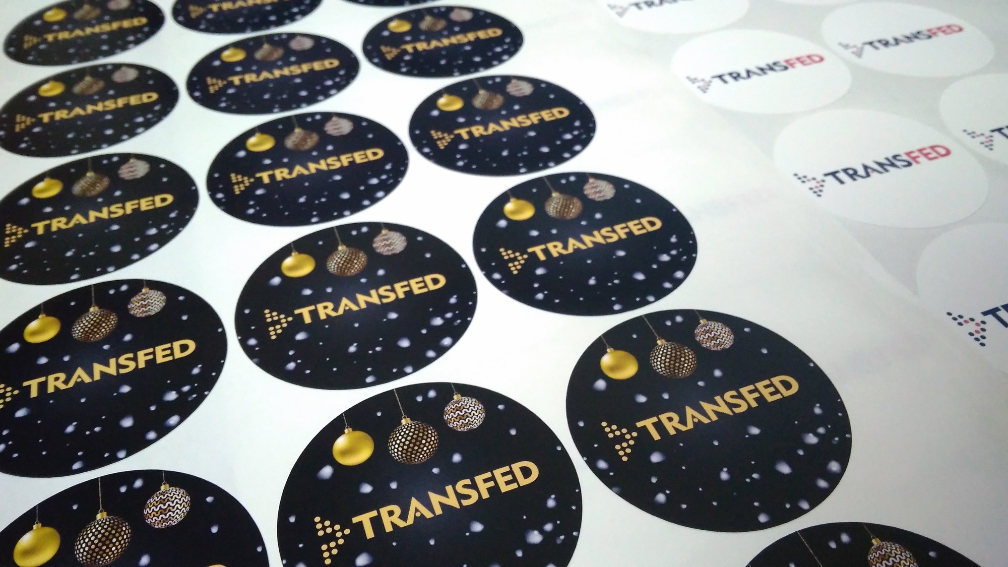 Arkusz okrągłych etykiet samoprzylepnych z logo 'TRANSFED' i motywem świątecznym (bombki i śnieżynki) na czarnym tle, część etykiet jest biała z logo.