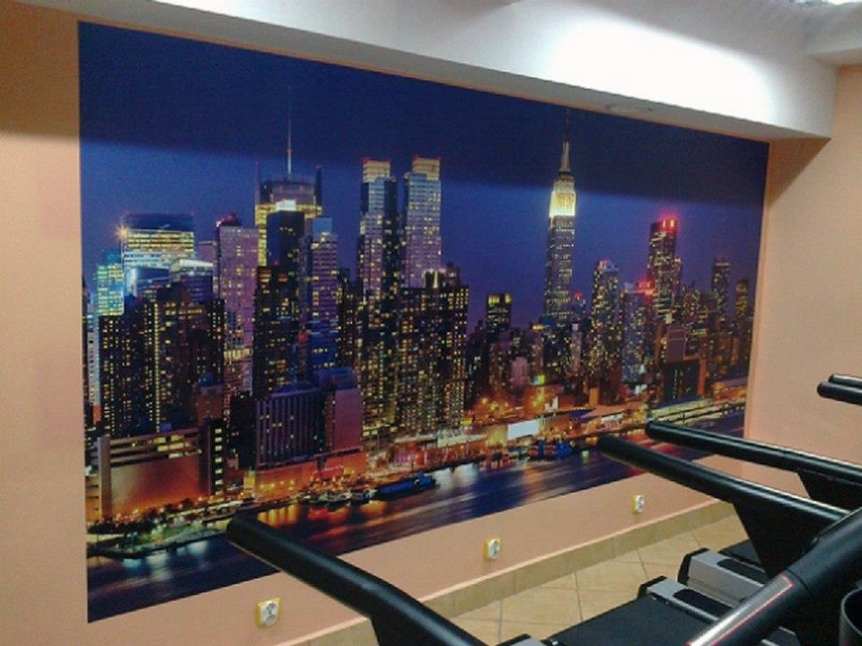 Tapeta z nocnym widokiem na panoramę Nowego Jorku, zamontowana na ścianie siłowni, widoczne bieżnie.