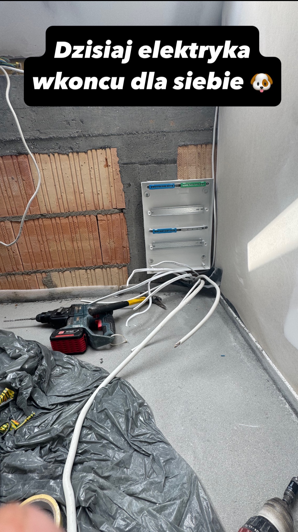 Rozdzielnia elektryczna w trakcie montażu w surowym wnętrzu. Widoczne kable, wiertarka i młotek. Ściana z cegły i surowy beton na podłodze.