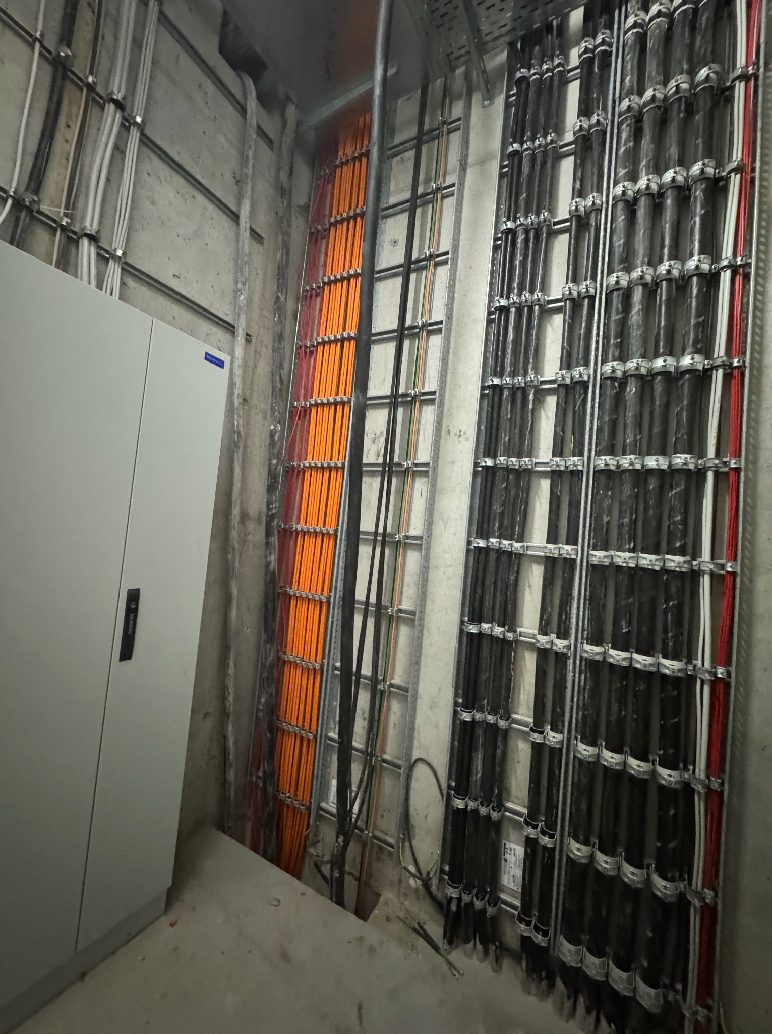 Rząd pomarańczowych kabli w metalowej drabince obok czarnych kabli w korytach, przy szarej szafie elektrycznej w surowym wnętrzu. Widok na instalacje.