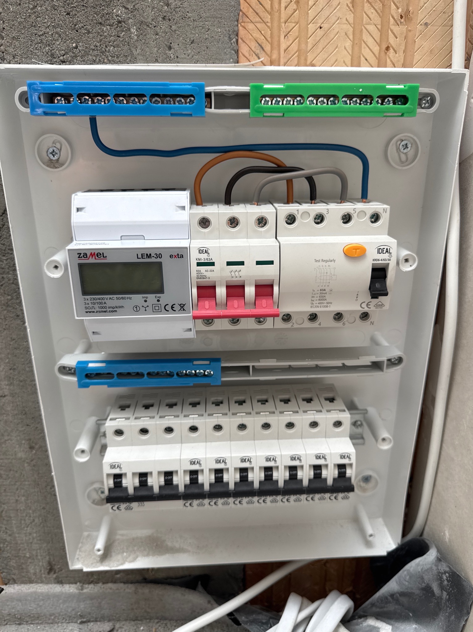Rozdzielnica elektryczna w białej obudowie z zamontowanym licznikiem Zamel LEM-30, bezpiecznikami IDEAL i listwami zaciskowymi w kolorach niebieskim i zielonym.