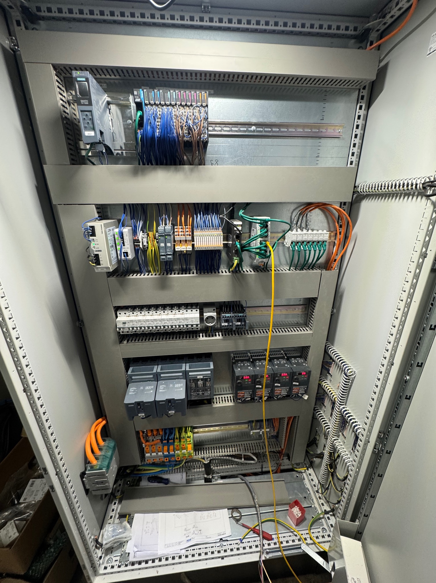 Rozdzielnica elektryczna w szarej obudowie z widocznymi przewodami, bezpiecznikami i elementami sterującymi. Widoczne schematy i narzędzia.