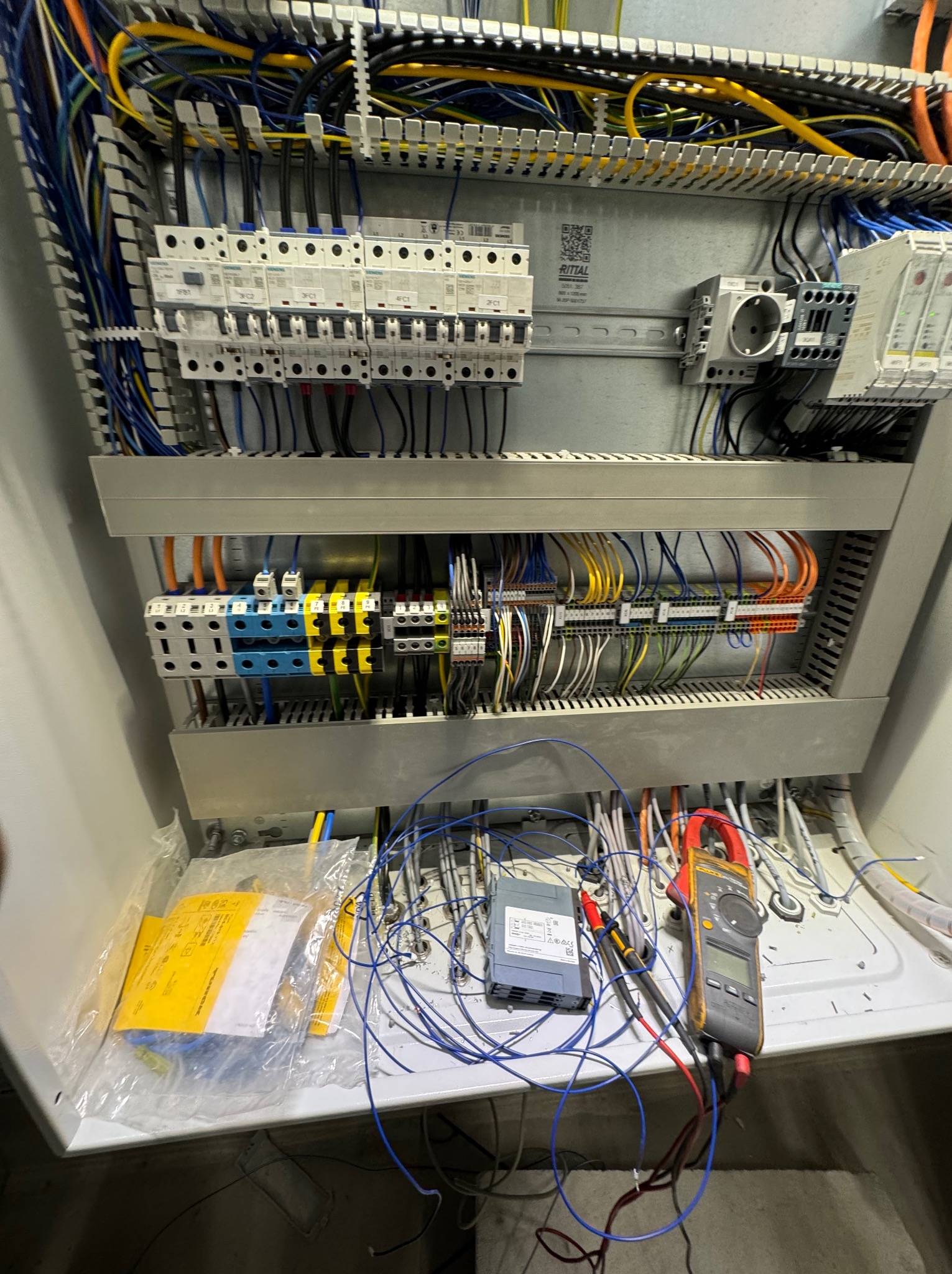 Otwarta szafa elektryczna z widocznymi bezpiecznikami, przewodami i miernikiem cęgowym. Widoczne elementy instalacji elektrycznej wewnątrz obudowy.