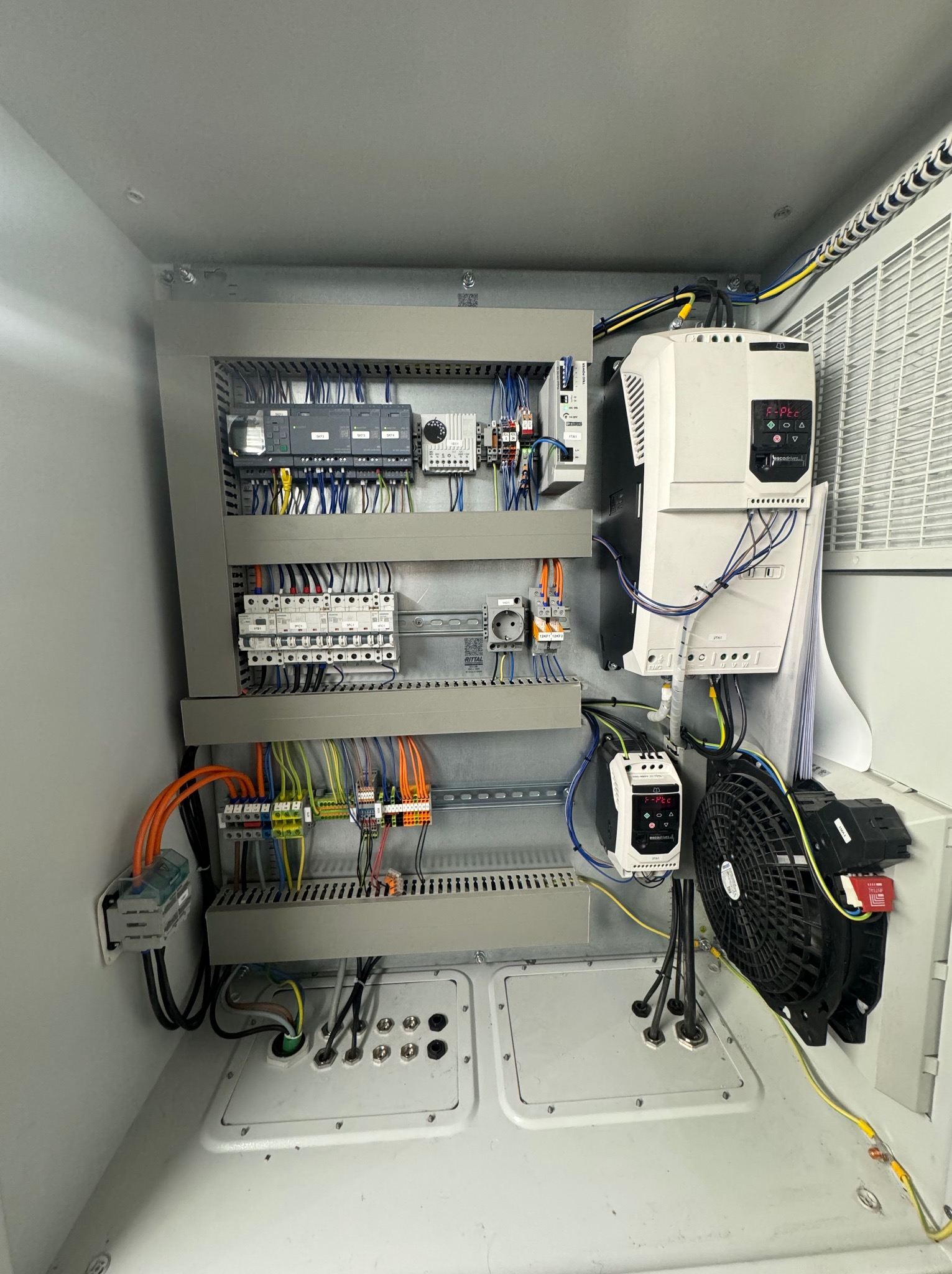 Otwarta szafa sterownicza z widocznymi elementami elektrycznymi: sterowniki PLC, bezpieczniki, okablowanie w kolorowych izolacjach, wentylator chłodzący.