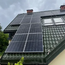 Panele słoneczne zamontowane na zielonym dachu z blachodachówki, widoczne okna dachowe i komin, pochmurne niebo.