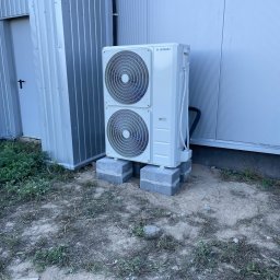 Sunsat - Zewnętrzna jednostka klimatyzacji marki Kaisai, umieszczona na betonowych blokach przy metalowym budynku z widocznymi przewodami.