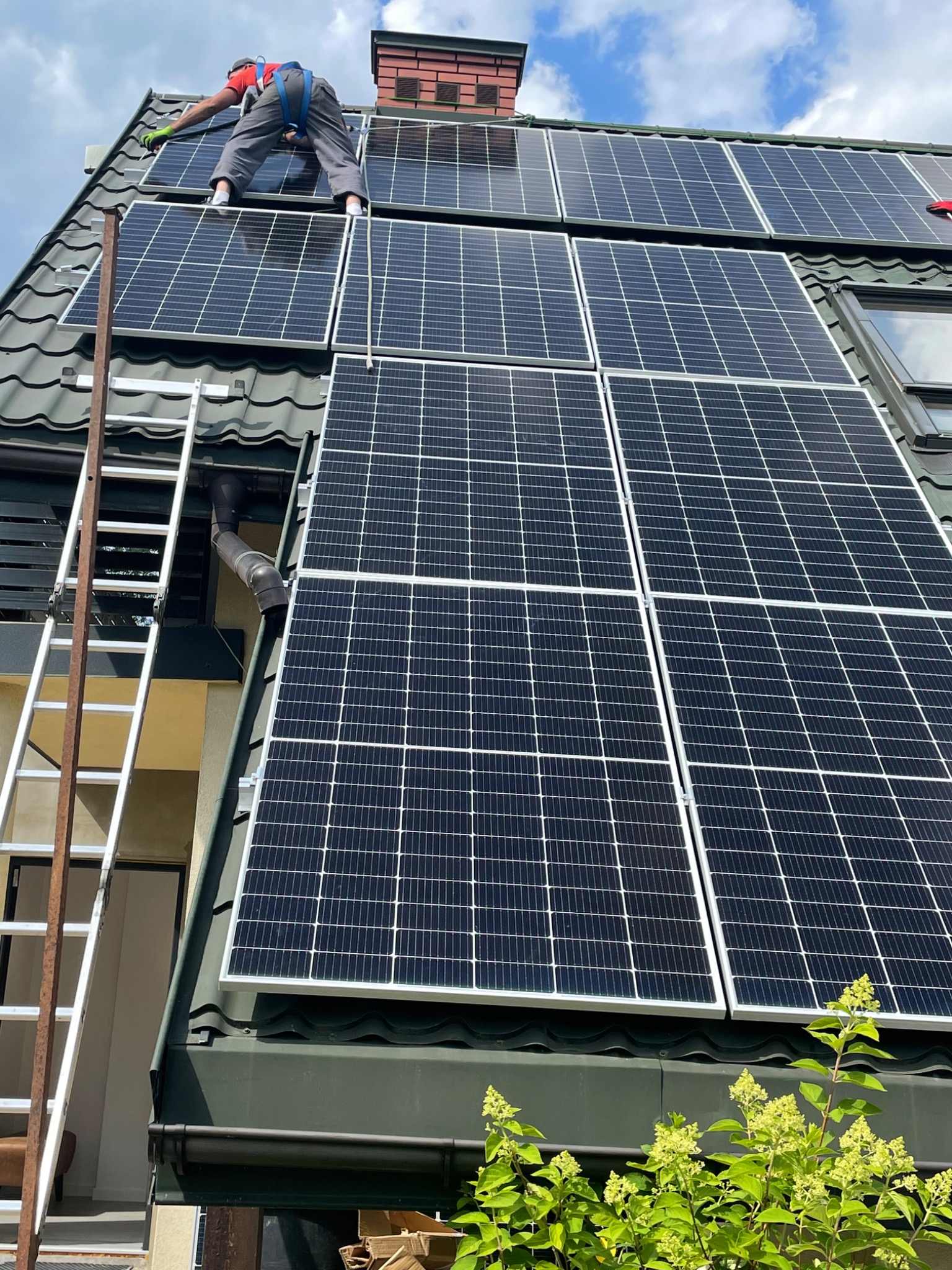 Instalator w czerwonej koszulce i szarych spodniach, zabezpieczony uprzężą, montuje panele słoneczne na dachu domu z ciemnozieloną dachówką, widoczna drabina oparta o rynnę.