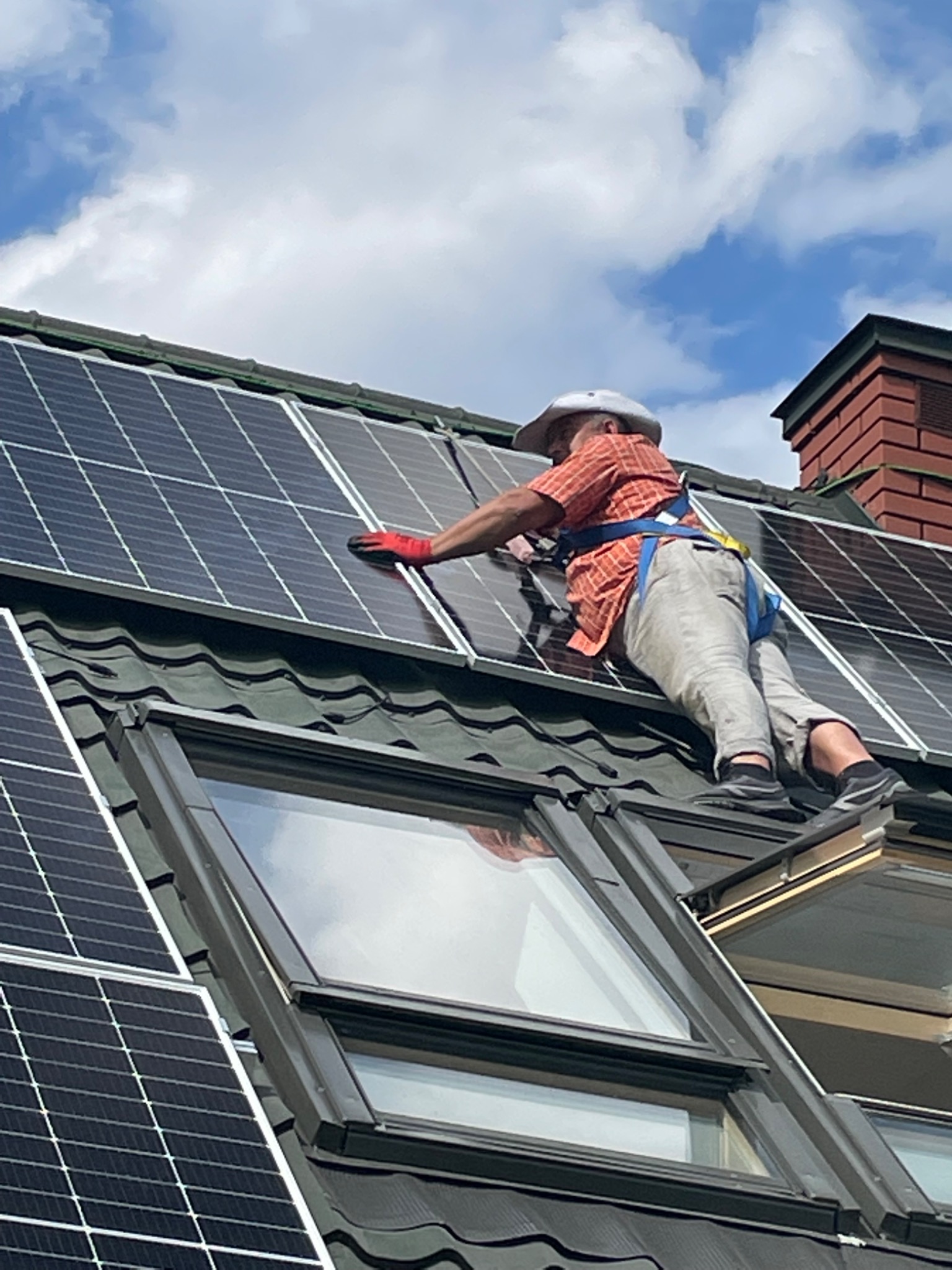 Pracownik w czerwonych rękawicach, z uprzężą i kapeluszu, montuje panele słoneczne na dachu z oknem dachowym, widoczne niebieskie niebo z chmurami.
