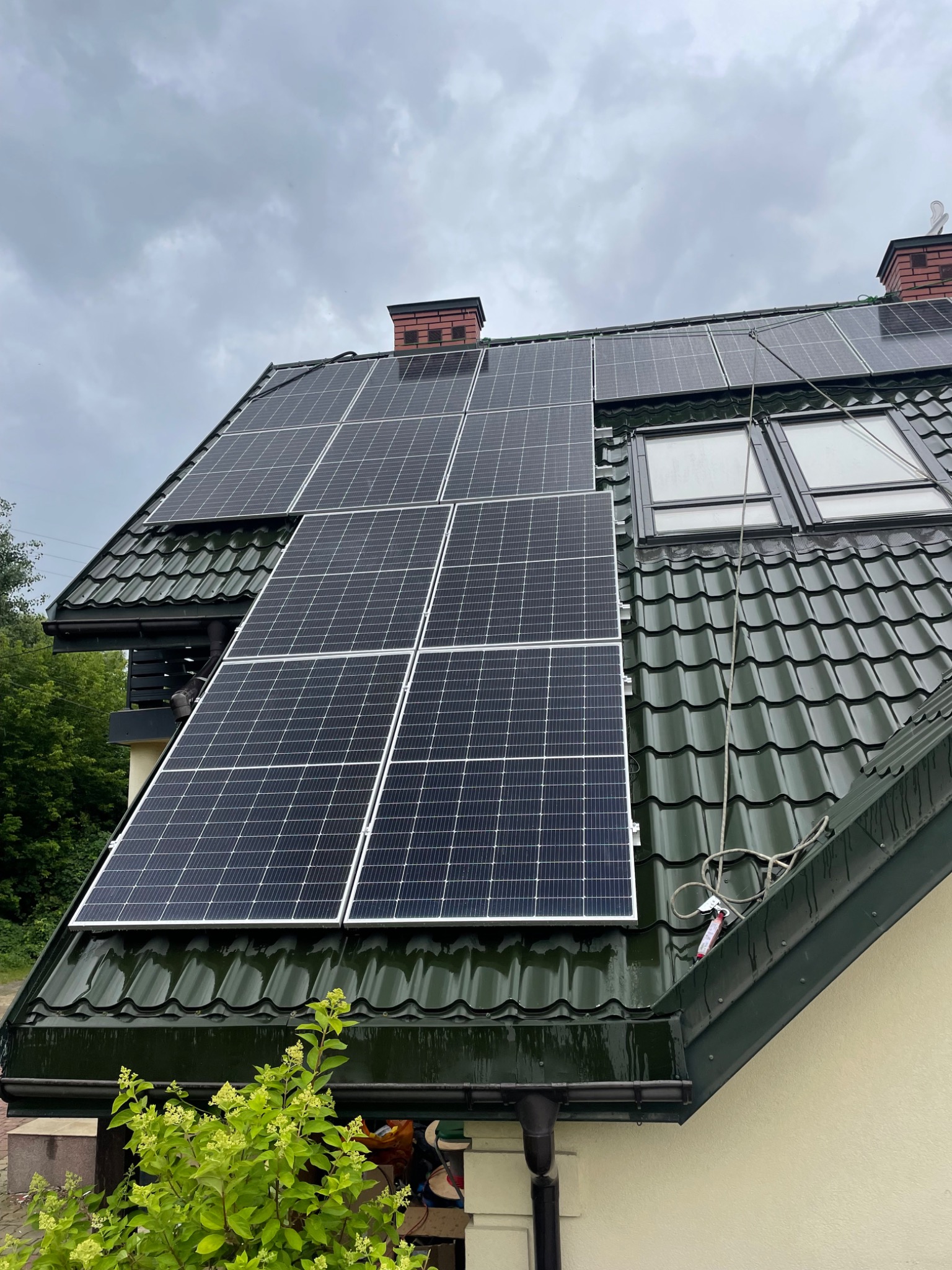 Panele słoneczne zamontowane na zielonym dachu z blachodachówki, widoczne okna dachowe i komin, pochmurne niebo.