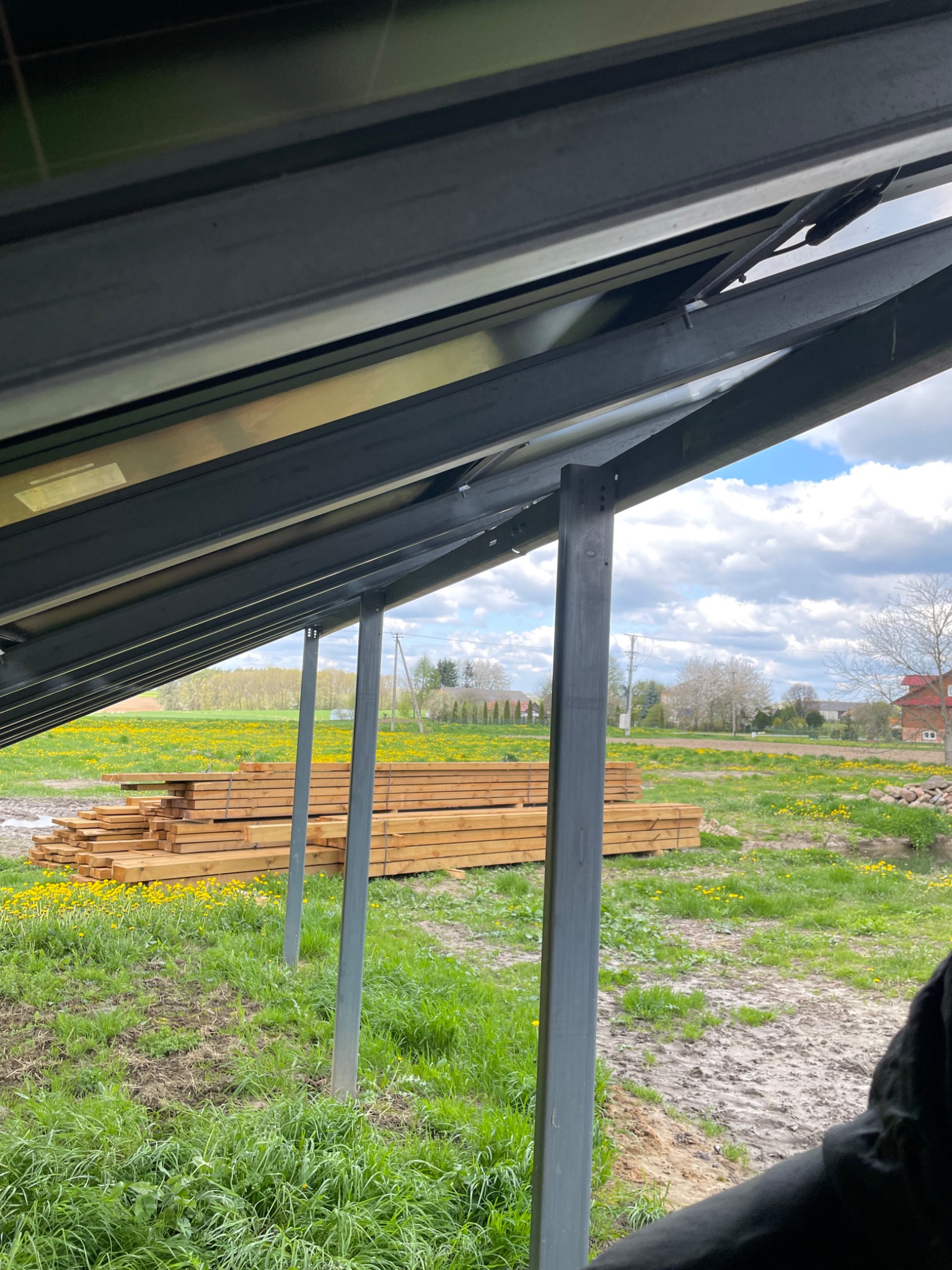 Widok spod konstrukcji wsporczej paneli słonecznych na łąkę z kwitnącymi mniszkami lekarskimi i stertę drewnianych belek, w tle zabudowania wiejskie pod błękitnym niebem z chmurami.