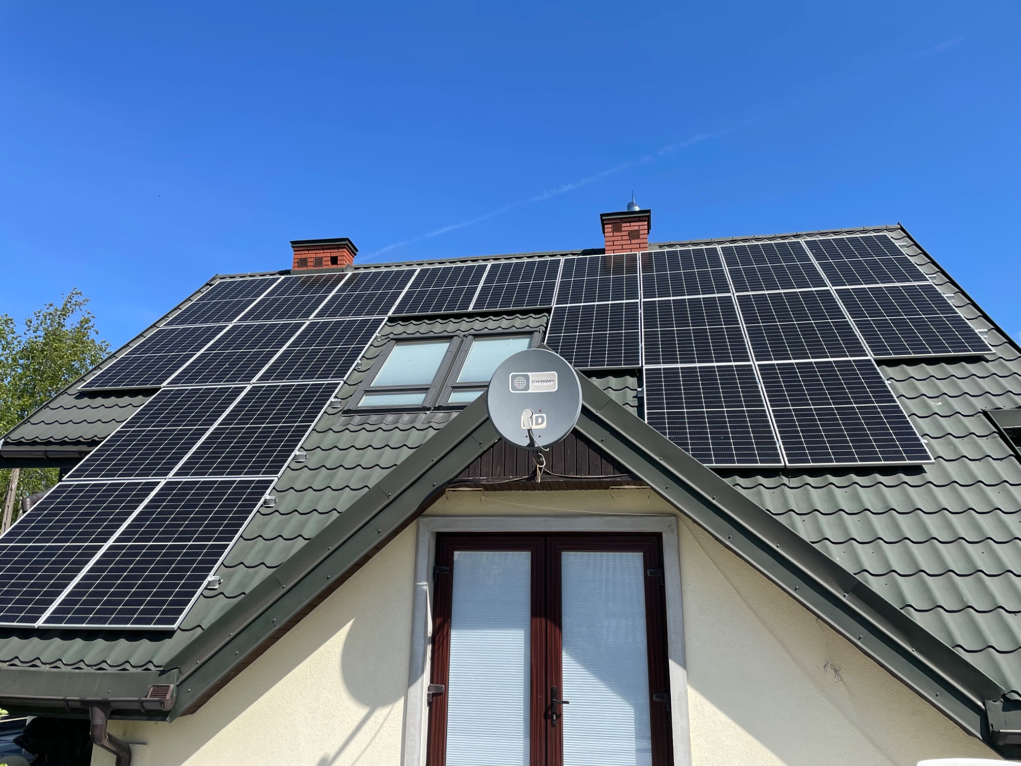 Panel fotowoltaiczny na dachu domu z dachówką w odcieniach szarości, widoczna antena satelitarna i okno dachowe, błękitne niebo w tle.