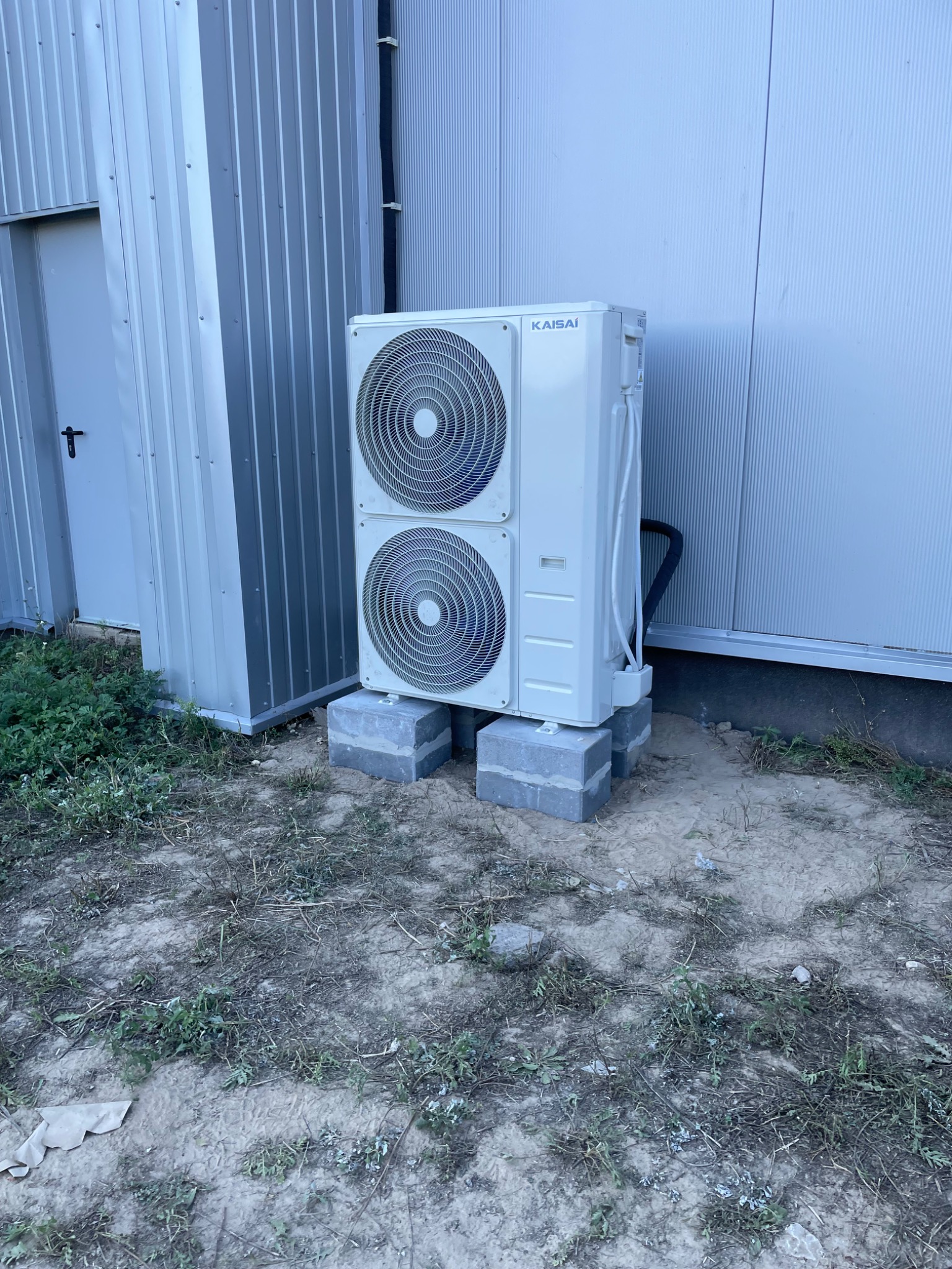 Zewnętrzna jednostka klimatyzacji marki Kaisai, umieszczona na betonowych blokach przy metalowym budynku z widocznymi przewodami.