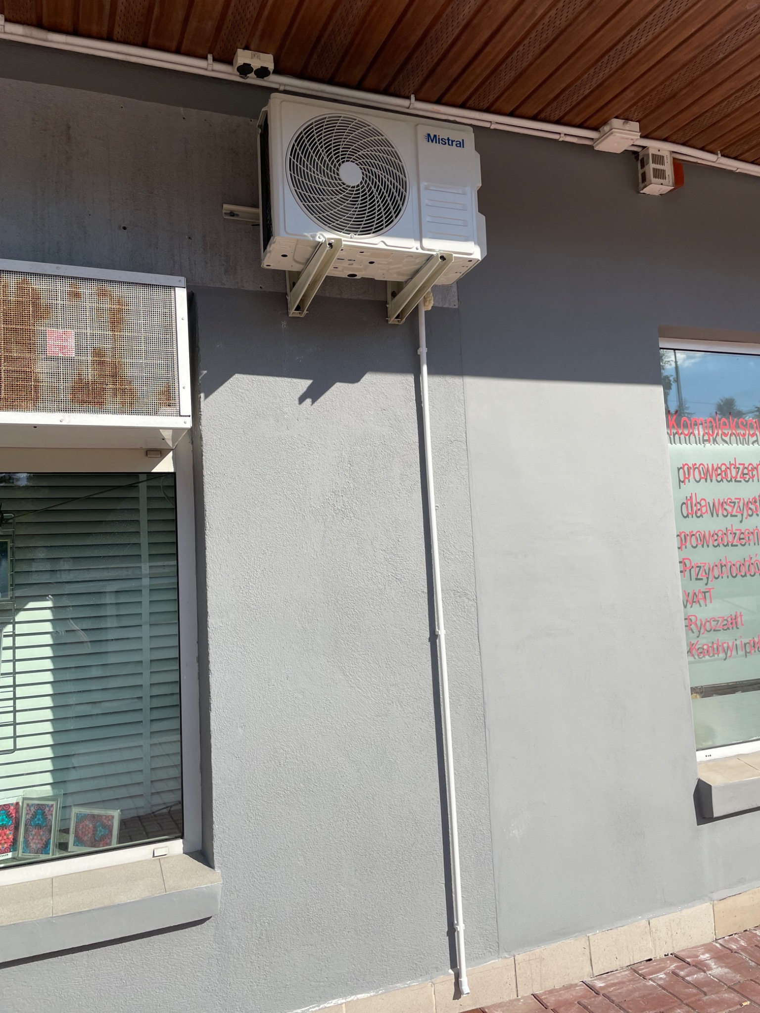 Zewnętrzna jednostka klimatyzacji Mistral zamontowana na szarej ścianie budynku, widoczna instalacja elektryczna, okno z żaluzjami i szyba z napisem reklamowym.