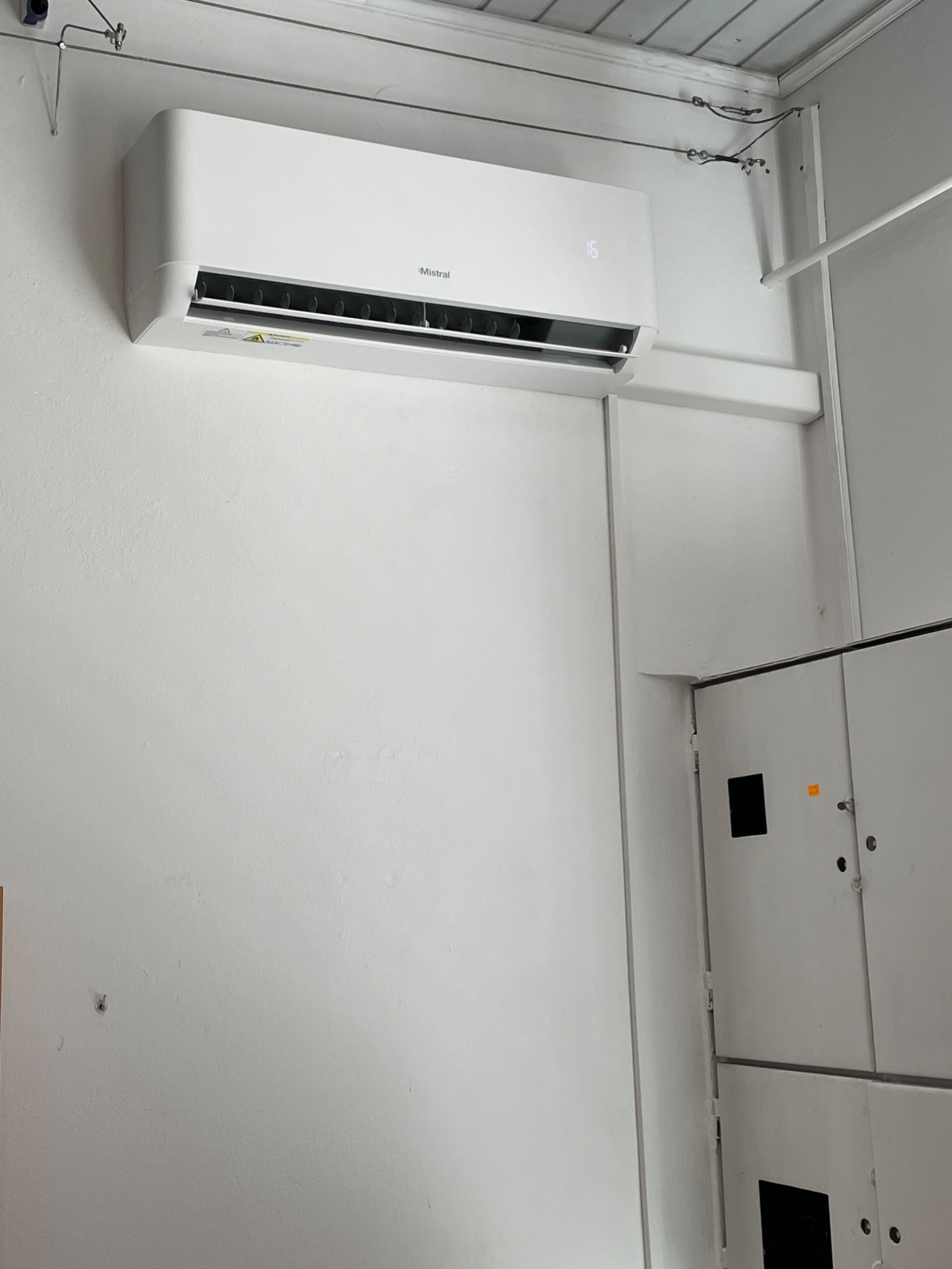 Biały klimatyzator marki Mistral zamontowany na białej ścianie w pomieszczeniu biurowym, obok rozdzielnicy elektrycznej.