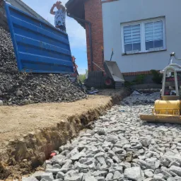 Przygotowanie terenu pod fundament: wykop, wysypany gruzem, ubijarka mechaniczna, na drugim planie kontener ze żwirem i pracownik.