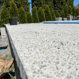 Szlifowana płyta betonowa z widoczną strukturą, ustawiona na zewnątrz, z zielonymi tujami w tle i elementami wyposażenia budowlanego, takimi jak wiadro i rura.