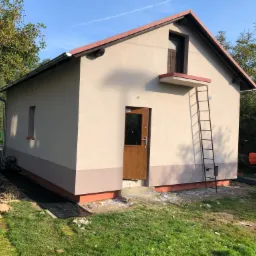 Otynkowany budynek gospodarczy z drabiną, świeżo po nałożeniu gładzi, z widocznymi śladami prac wykończeniowych wokół budynku.