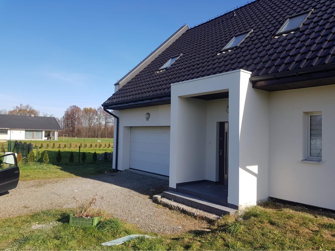 Nowoczesny dom jednorodzinny z białymi ścianami, czarną dachówką i garażem, widok z zewnątrz w słoneczny dzień.