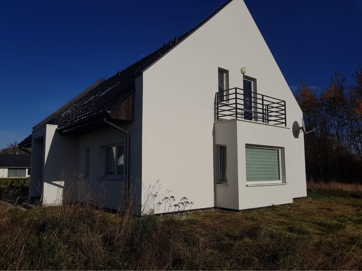 Elewacja nowo wybudowanego domu jednorodzinnego z balkonem i anteną satelitarną, widok z niskiej perspektywy na tle błękitnego nieba i drzew z jesiennymi liśćmi.