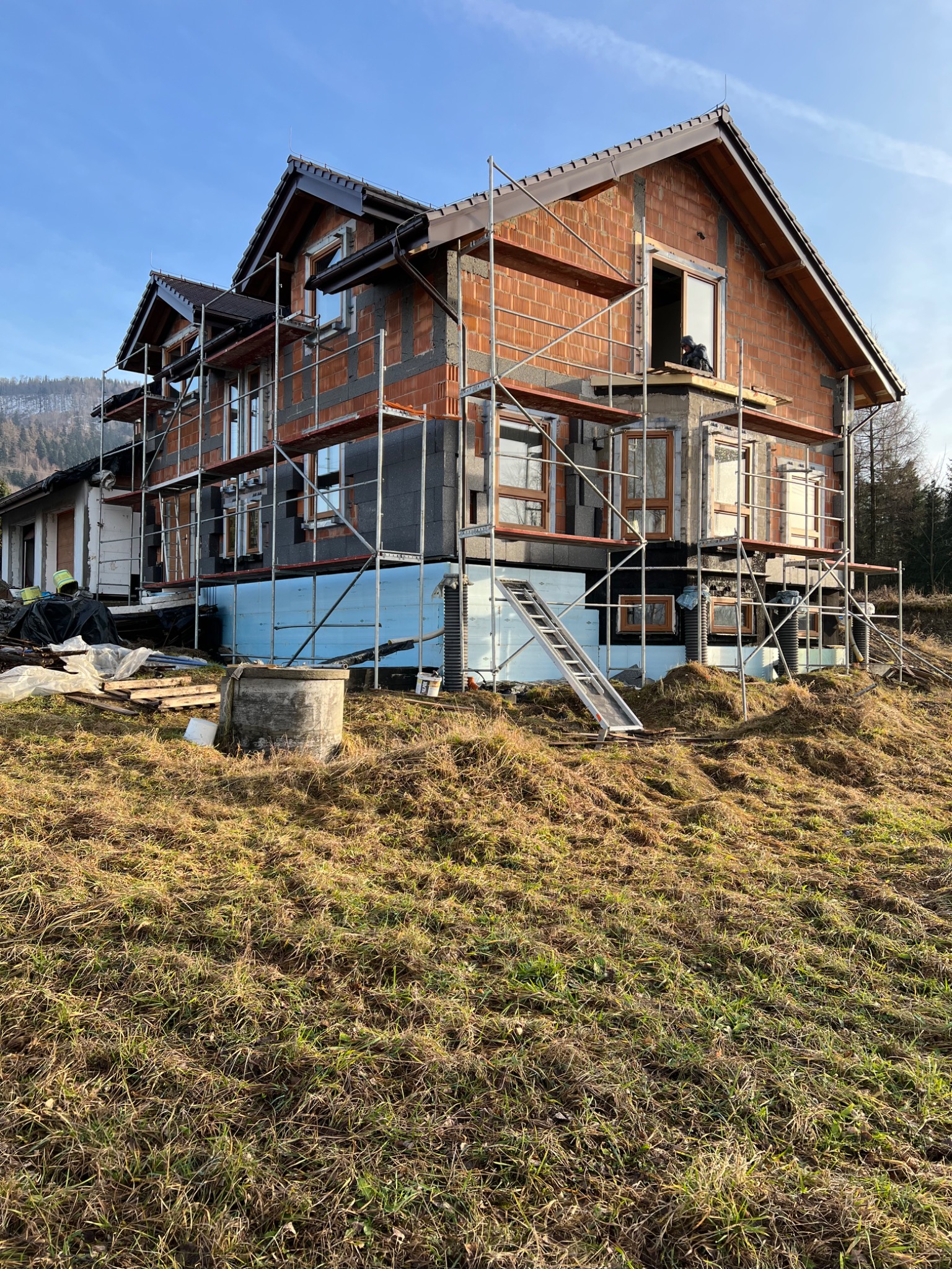 Dom w trakcie budowy: murowana konstrukcja z oknami, ocieplenie styropianem, rusztowania, widok na pagórkowaty teren z lasem w tle.