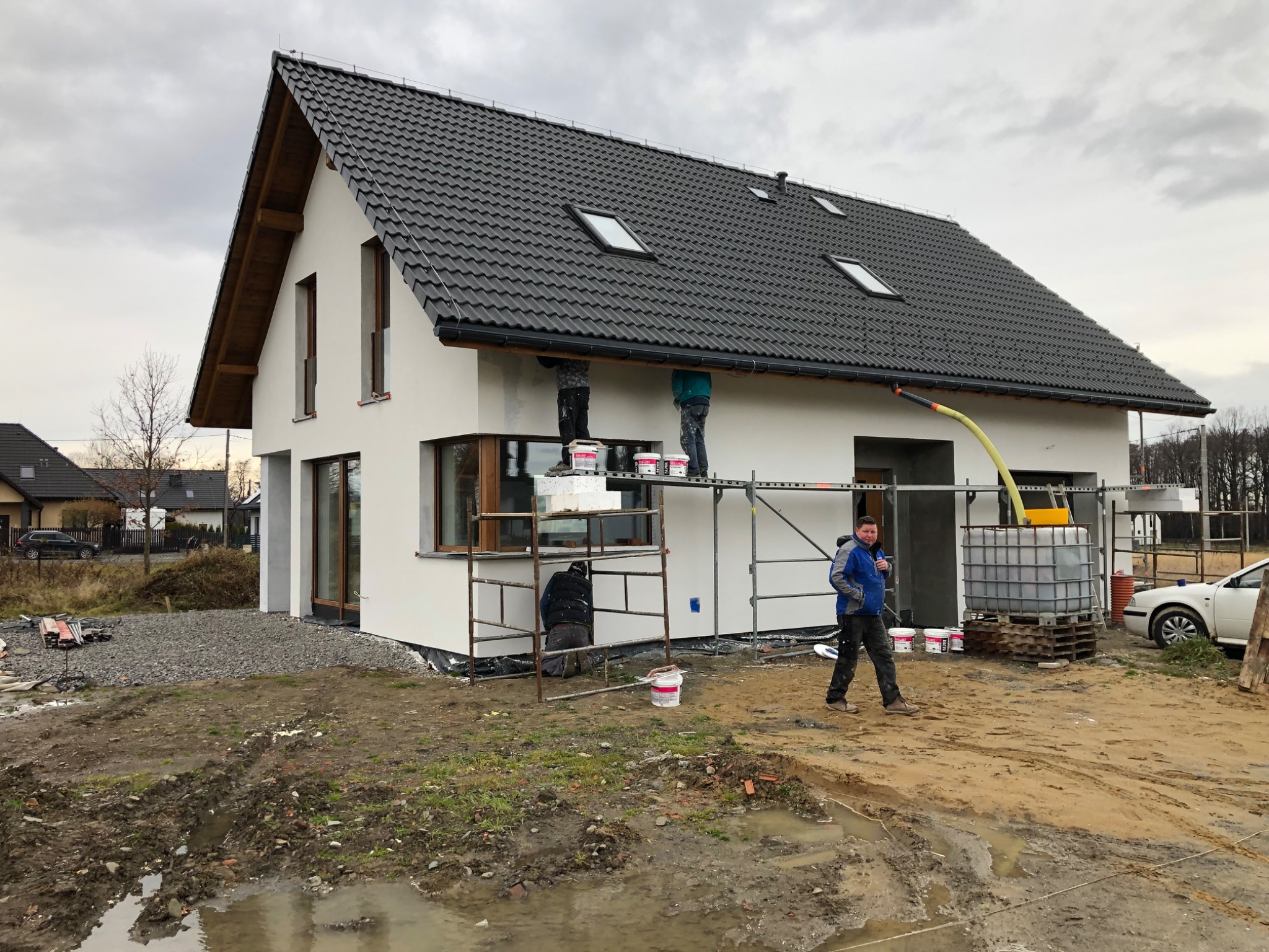 Dwóch pracowników na rusztowaniu aplikuje gładź na elewację domu jednorodzinnego z ciemną dachówką, jeden pracownik stoi na ziemi, widoczne wiadra z gładzią i żółty wąż do agregatu.