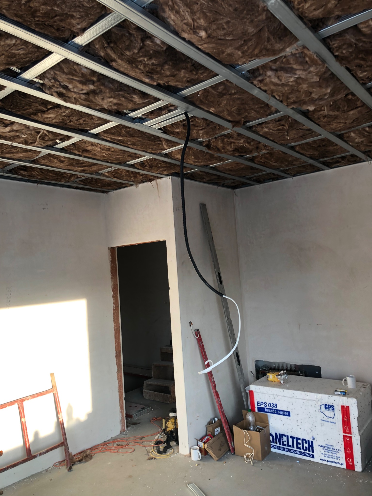 Wnętrze w trakcie prac wykończeniowych: widoczna konstrukcja sufitu podwieszanego z izolacją z wełny mineralnej, ściany przygotowane pod gładzie, narzędzia i materiały budowlane w pomieszczeniu.