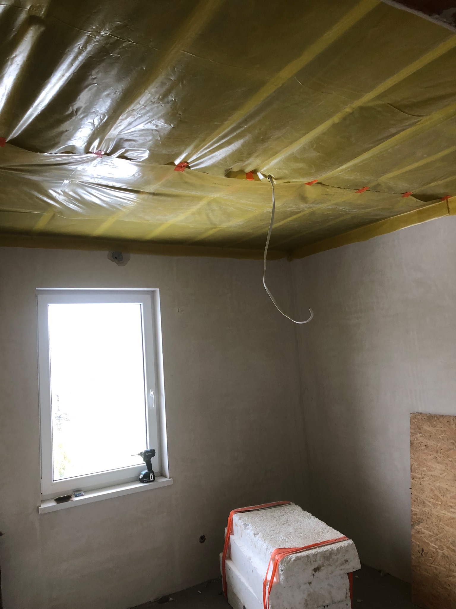 Wnętrze w trakcie prac wykończeniowych: sufit zabezpieczony folią z widocznymi mocowaniami, ściany po nałożeniu gładzi, okno z wkrętarką na parapecie, oraz styropian w pomarańczowej folii w rogu...
