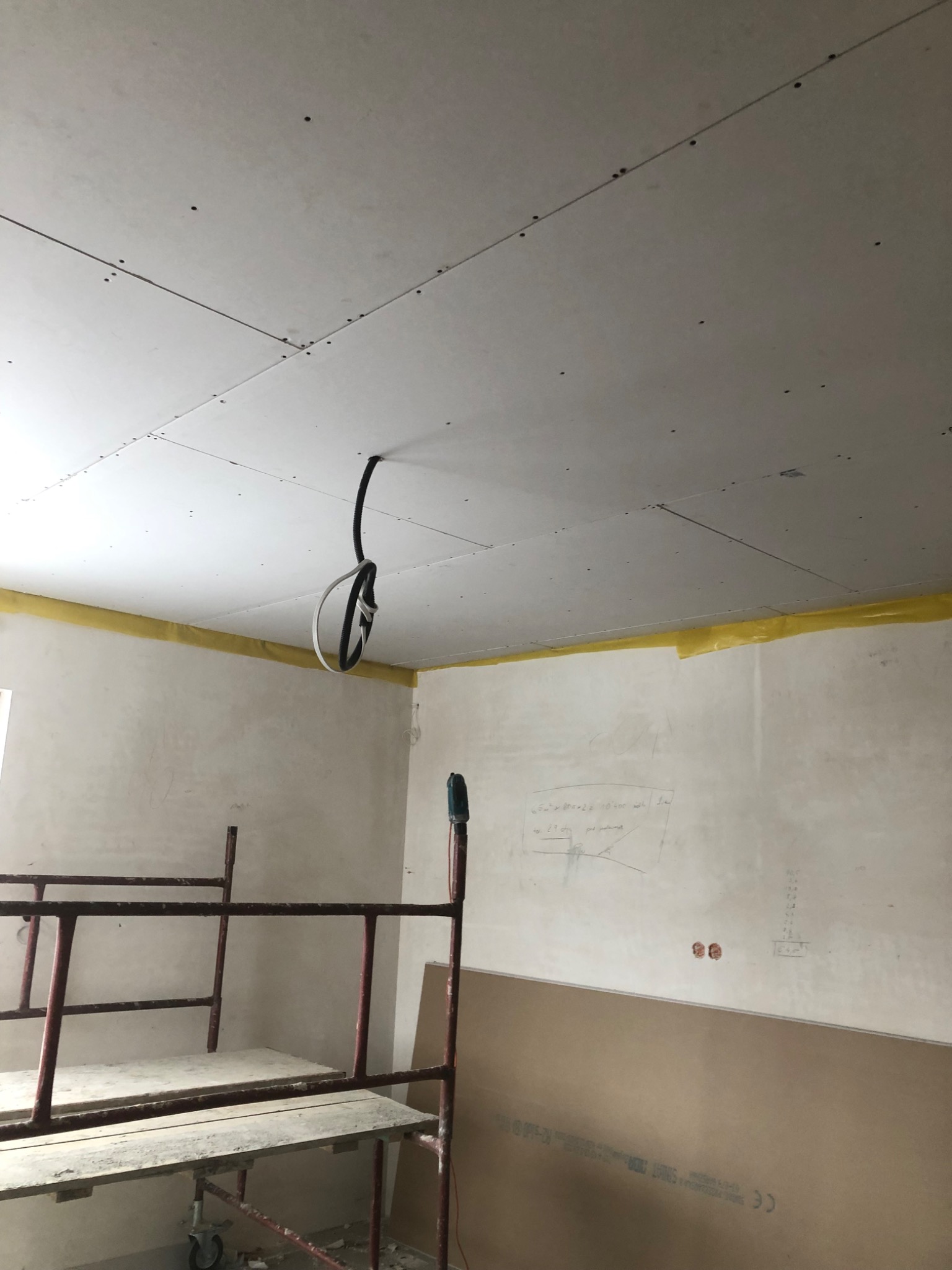 Montaż sufitu z płyt kartonowo-gipsowych: widoczne płyty przytwierdzone do konstrukcji, wystające przewody elektryczne, ślady ołówka na ścianie i fragment rusztowania.