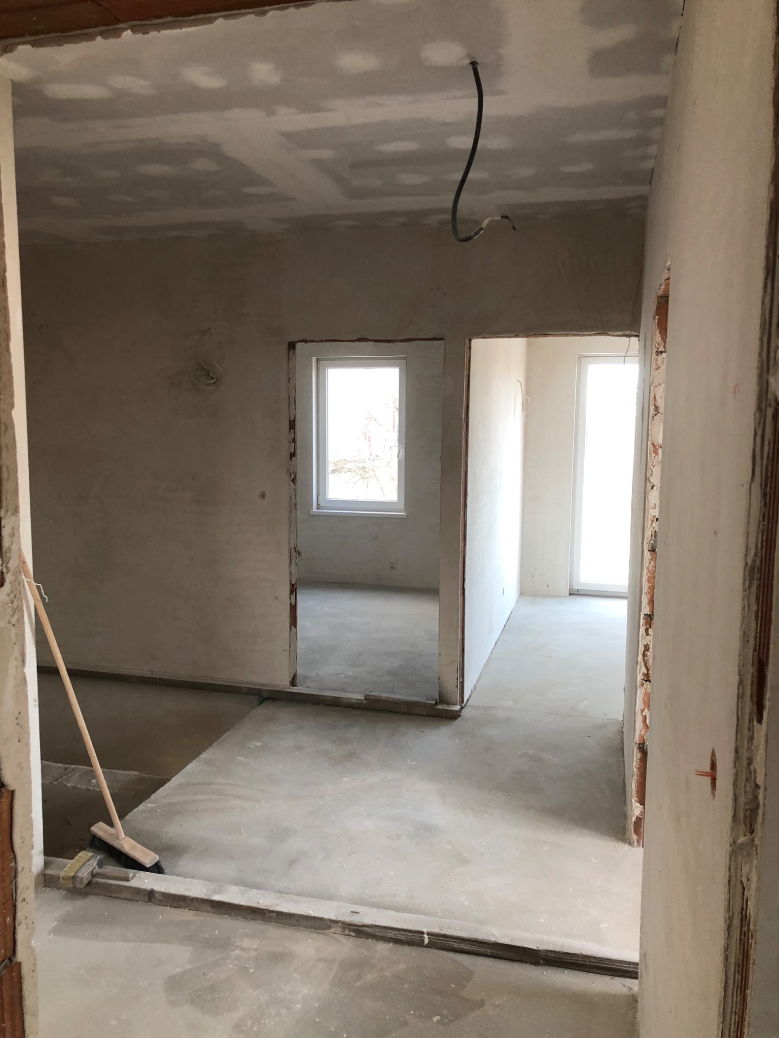 Wnętrze w trakcie prac wykończeniowych: surowe ściany, gładzie na suficie, wystający przewód elektryczny, przygotowany podkład podłogowy, widoczna miotła oparta o ścianę.