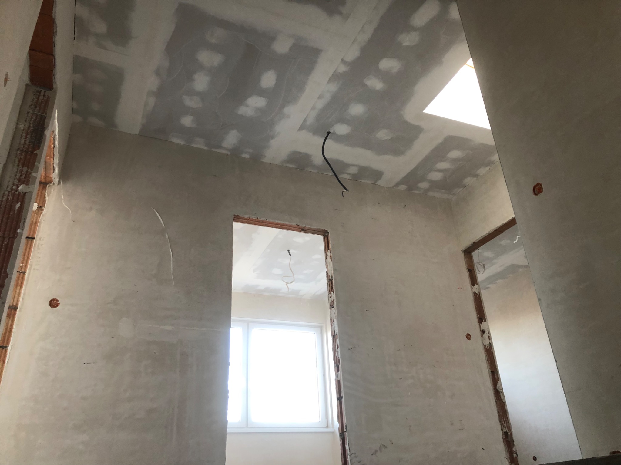 Wnętrze w trakcie prac wykończeniowych: surowa ściana z widocznymi punktami elektrycznymi, częściowo wykończony sufit z płyt kartonowo-gipsowych z widocznym przewodem elektrycznym, oraz widok...