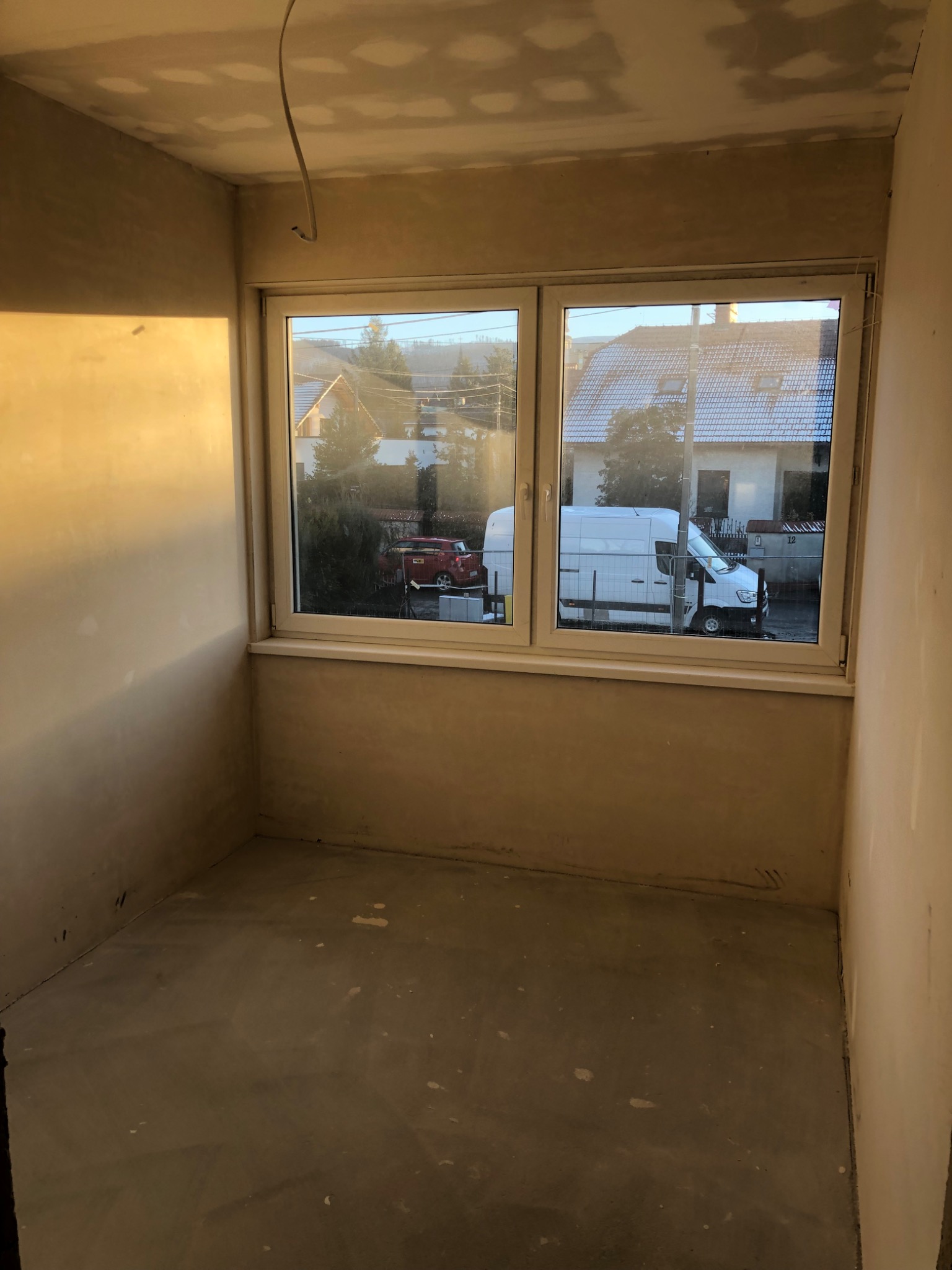 Pokój w trakcie prac przygotowawczych: ściany pokryte warstwą wyrównującą, widoczne przewody elektryczne, okno z widokiem na ulicę i zaparkowany biały samochód dostawczy.