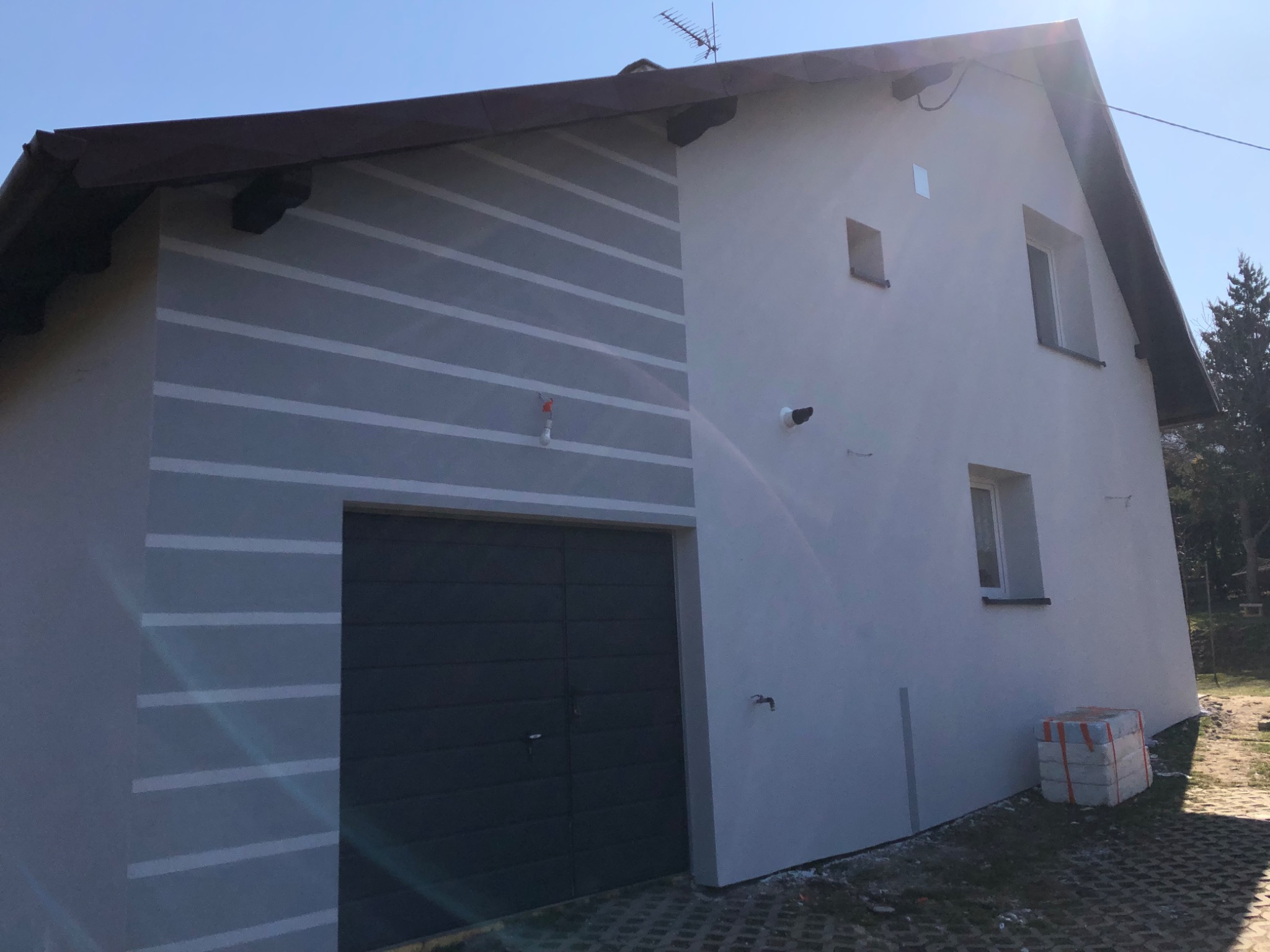 Świeżo otynkowany dom z garażem, z dekoracyjnymi pasami na elewacji, widoczne materiały budowlane obok budynku.