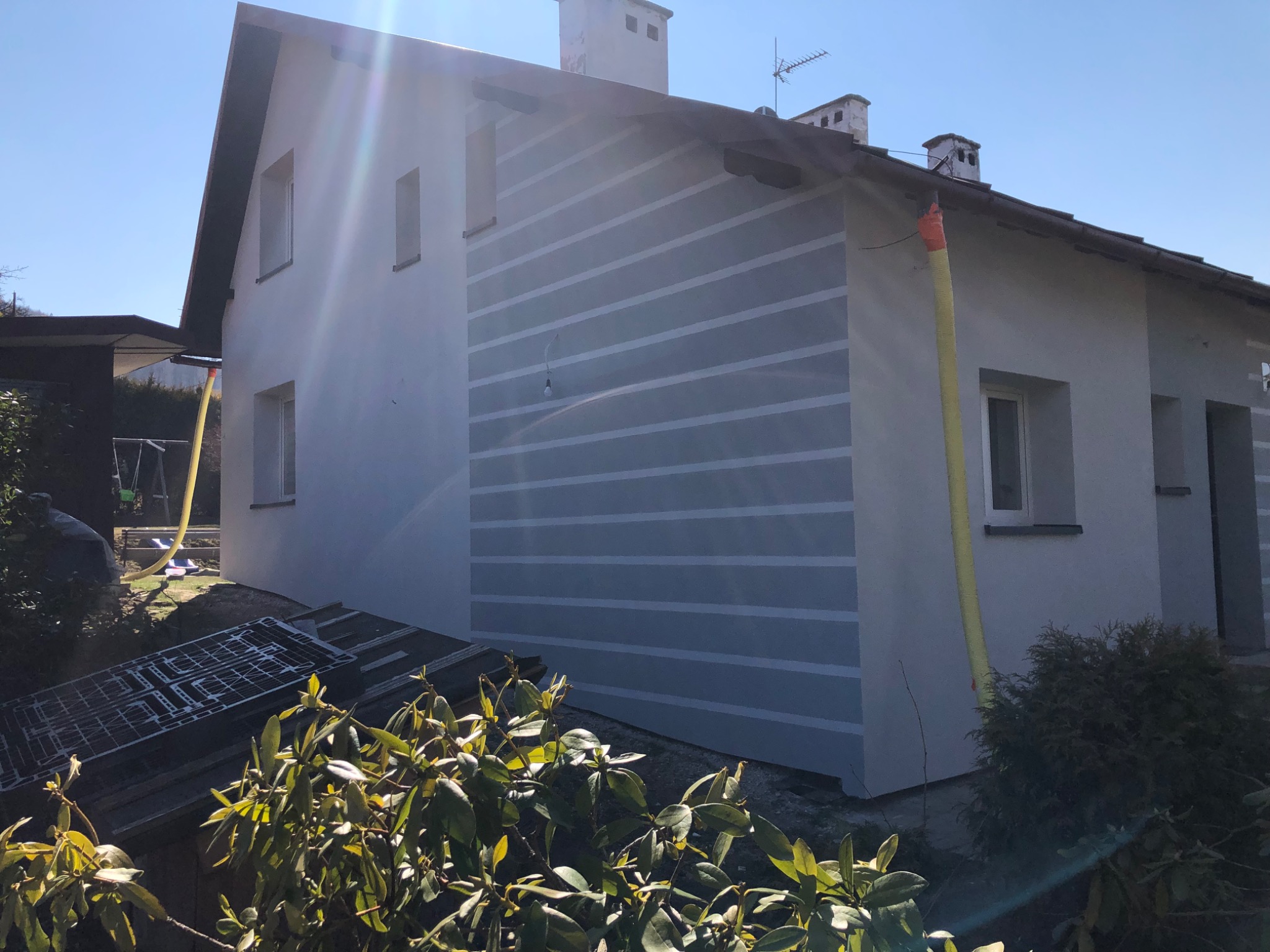 Elewacja domu jednorodzinnego z poziomymi pasami w trakcie malowania, widoczne rusztowania i żółty przewód wentylacyjny.