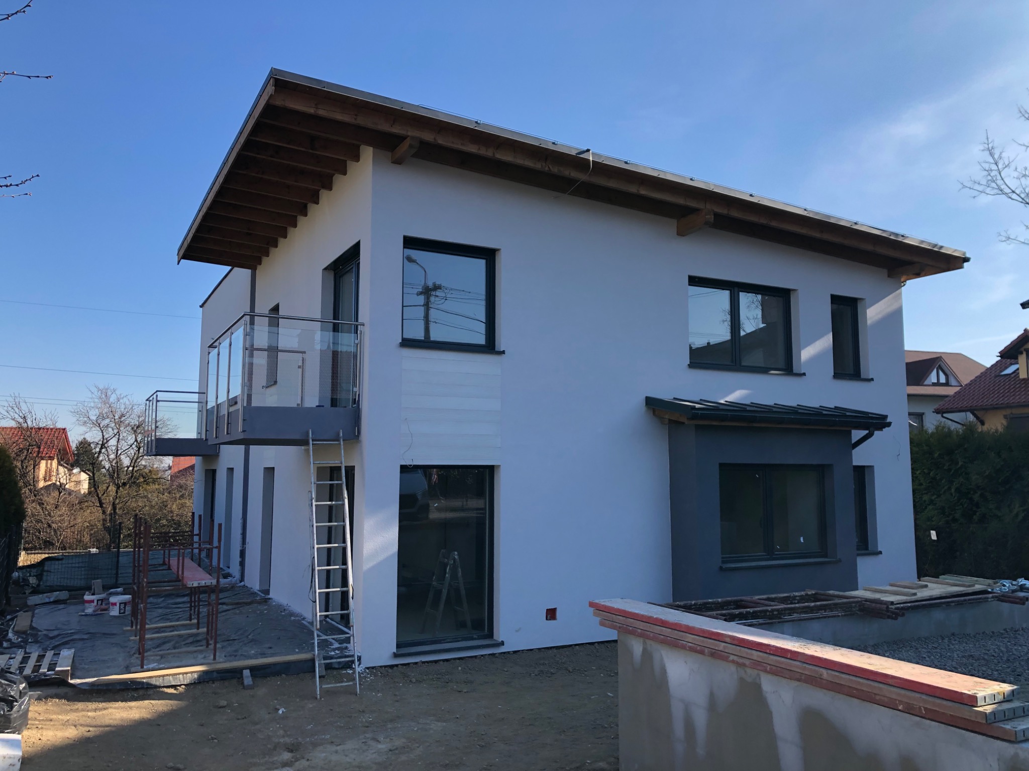 Nowy dom jednorodzinny z białymi ścianami zewnętrznymi, szarymi akcentami i balkonem z przezroczystą balustradą, widoczny na etapie wykończenia, ze stojącą drabiną i materiałami budowlanymi w pobliżu