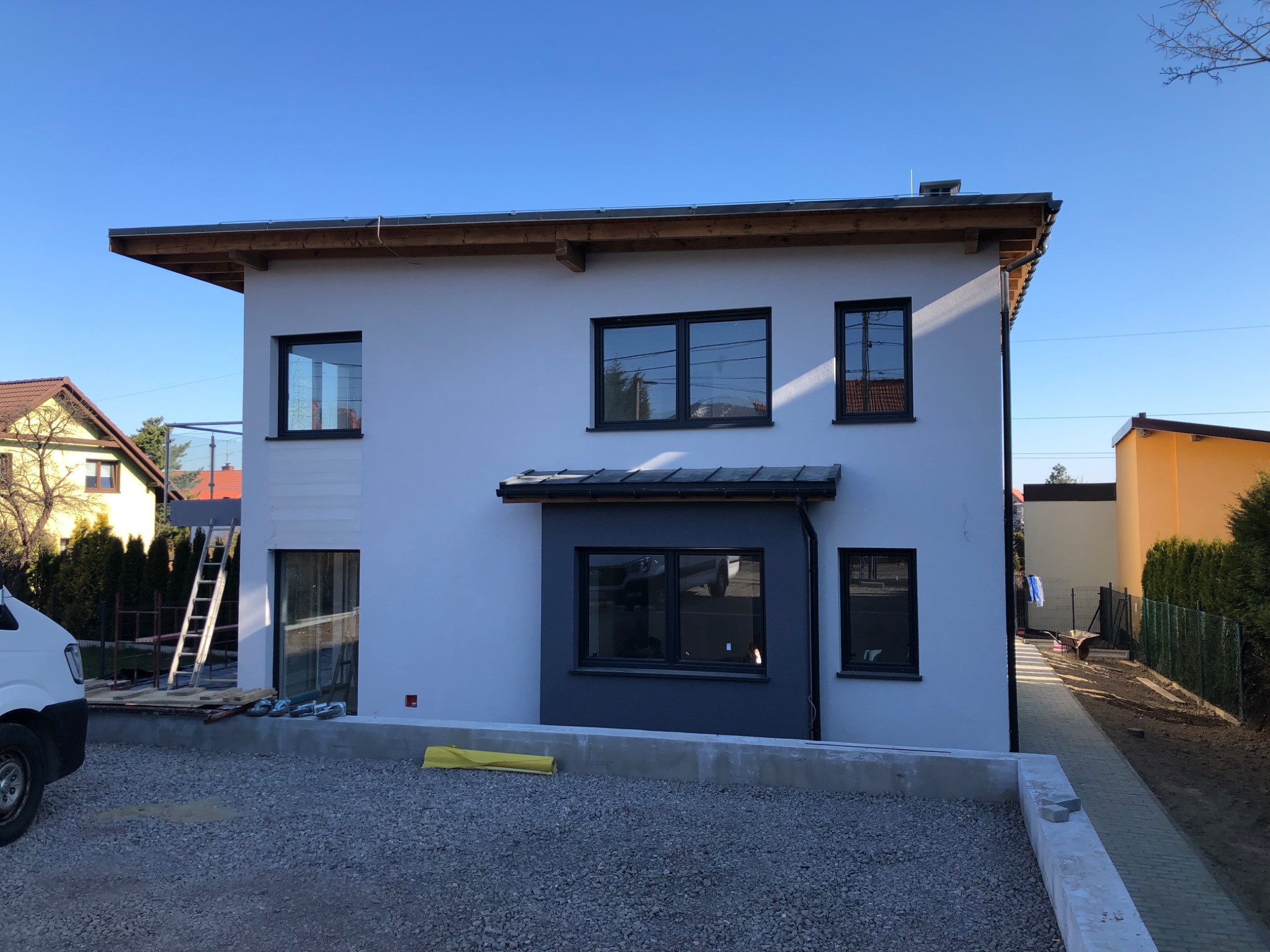 Dwukondygnacyjny dom jednorodzinny z białymi ścianami i ciemnymi oknami, widoczny z zewnątrz podczas prac wykończeniowych, z gruzem na podwórku i drabiną opartą o ścianę.