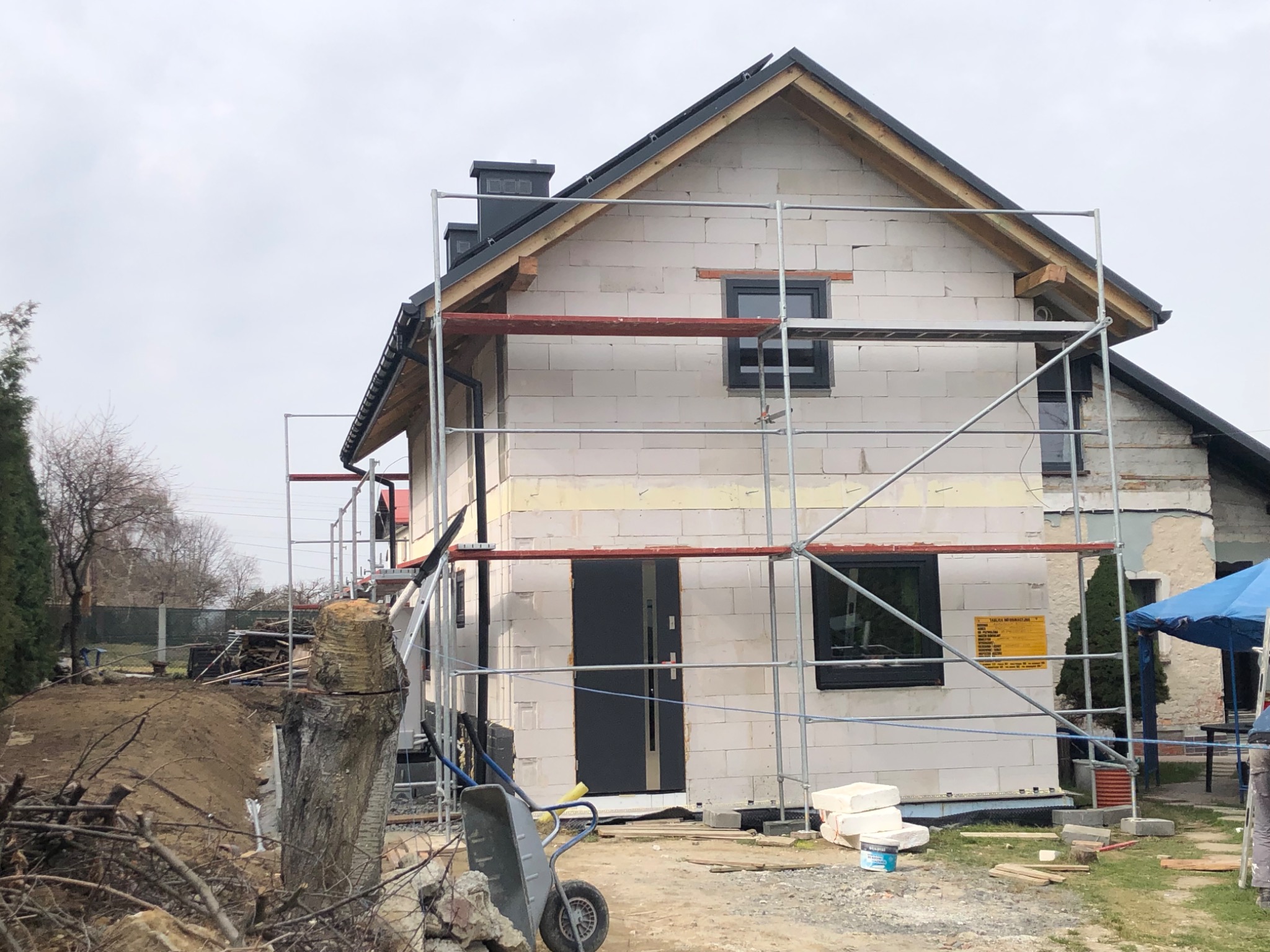 Budynek w trakcie budowy z białych bloczków, widoczne rusztowanie, zamontowane okna i drzwi, na ścianie żółta tablica informacyjna, w tle drzewa i niebo.