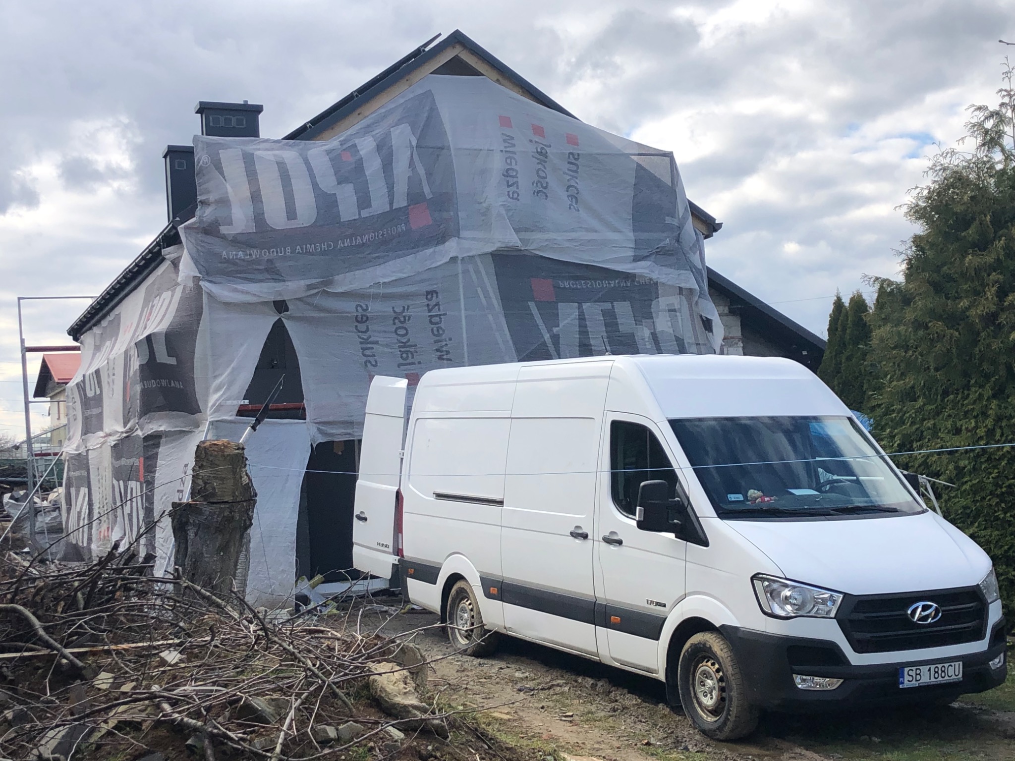 Dom w trakcie remontu elewacji, zabezpieczony folią budowlaną, biały van zaparkowany przed budynkiem, widoczne gałęzie i gruz.