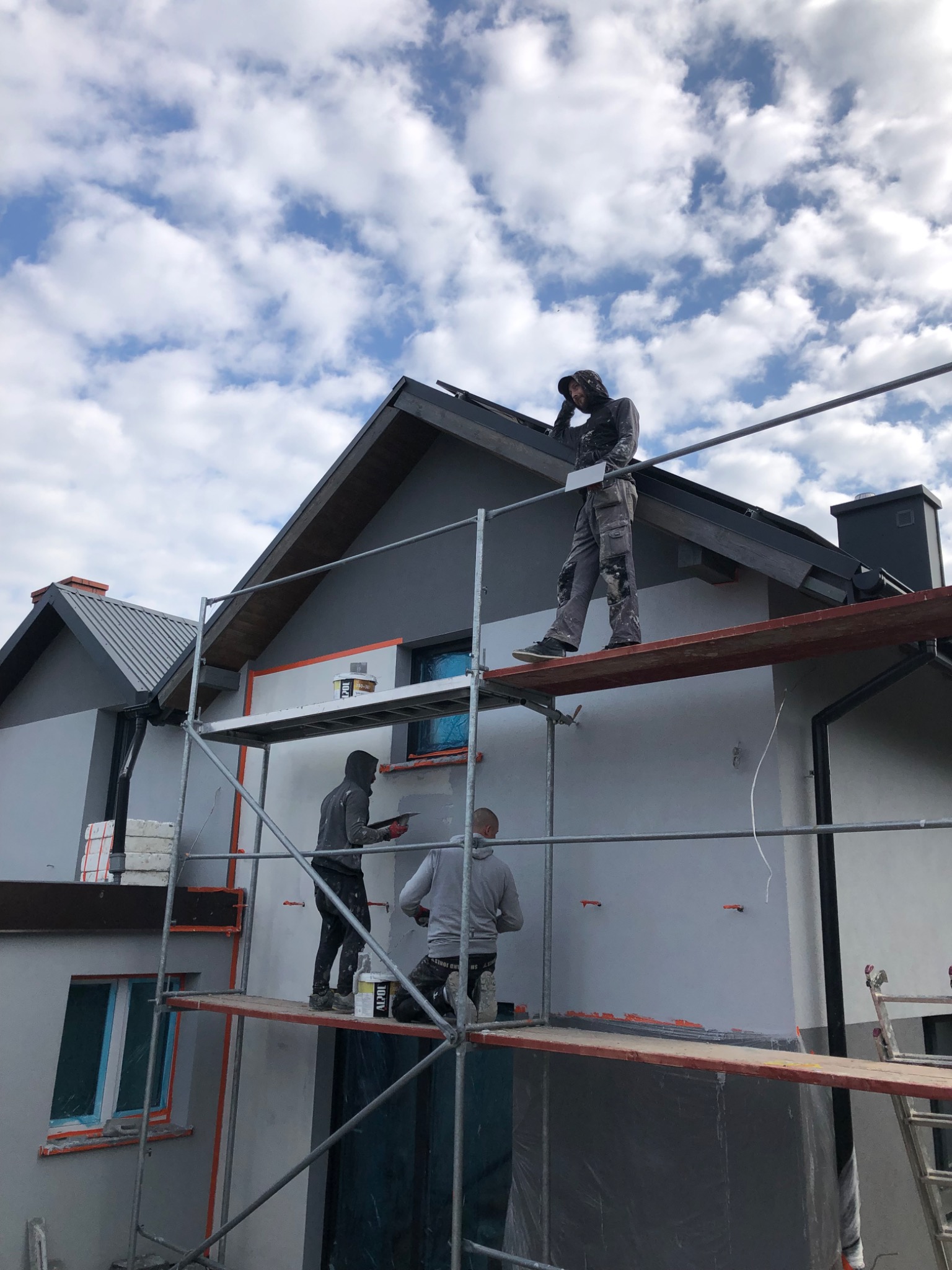 Trzech mężczyzn na rusztowaniu podczas nakładania gładzi na elewację dwukondygnacyjnego domu z szarą dachówką, widok z dołu na błękitne niebo z chmurami.