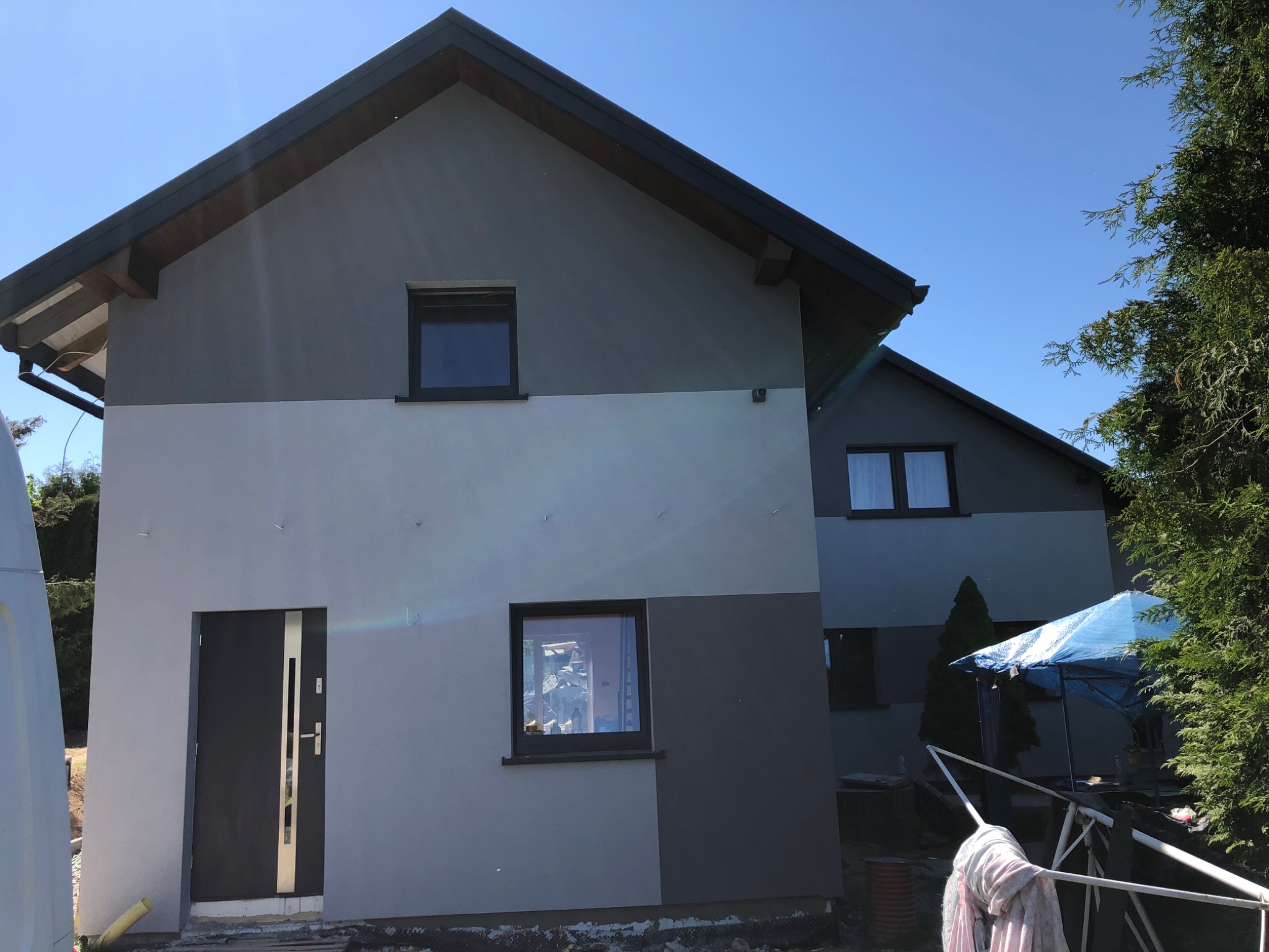 Dwukondygnacyjny dom jednorodzinny z szarą fasadą podzieloną na dwa odcienie, widok z zewnątrz, z boku widoczny fragment niebieskiej plandeki i zieleń drzew.