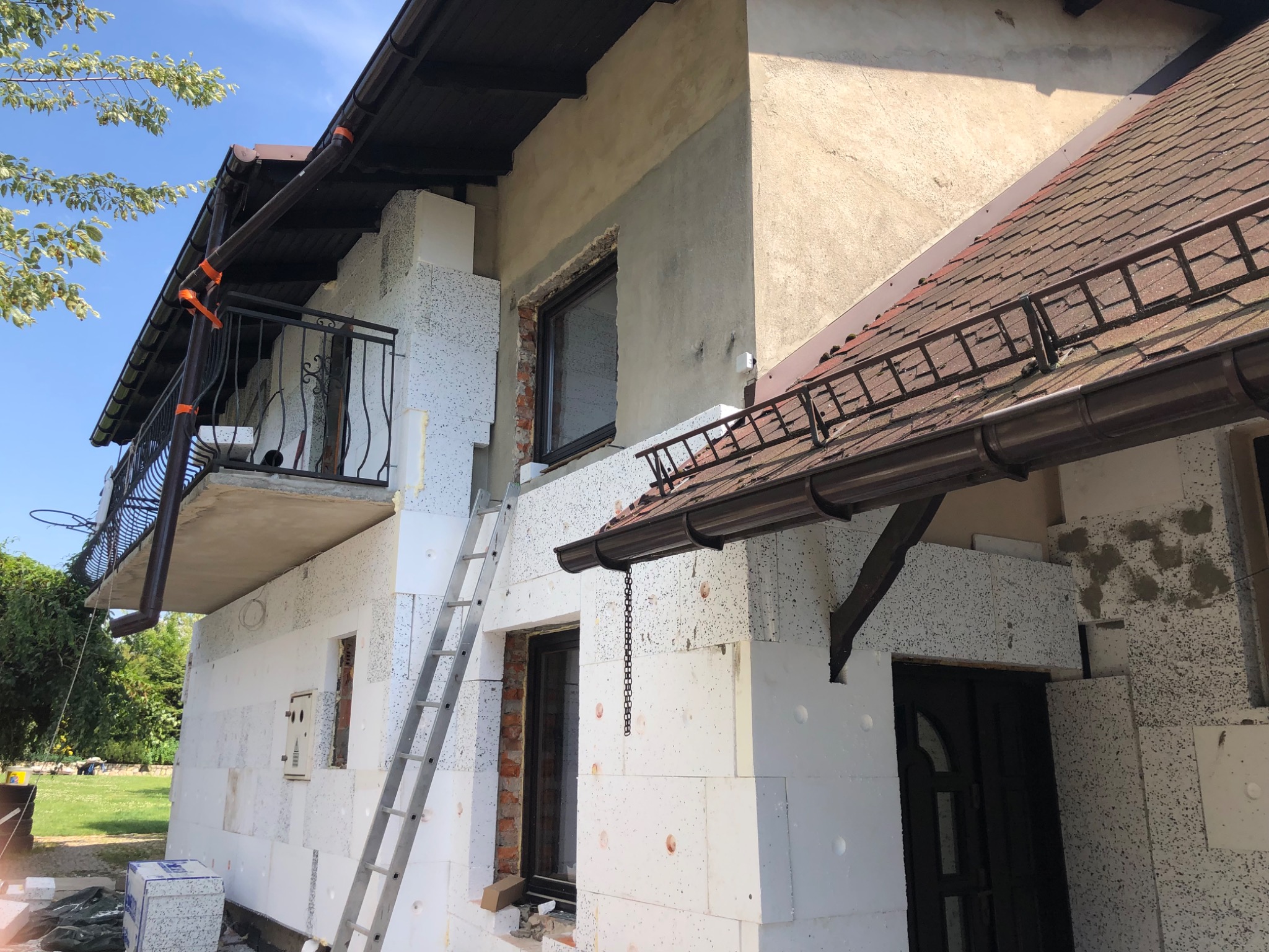 Ocieplanie budynku styropianem, widoczne płyty styropianowe na elewacji, drabina oparta o ścianę, przygotowane okna do montażu, częściowo otynkowana ściana, balkon z czarną balustradą.
