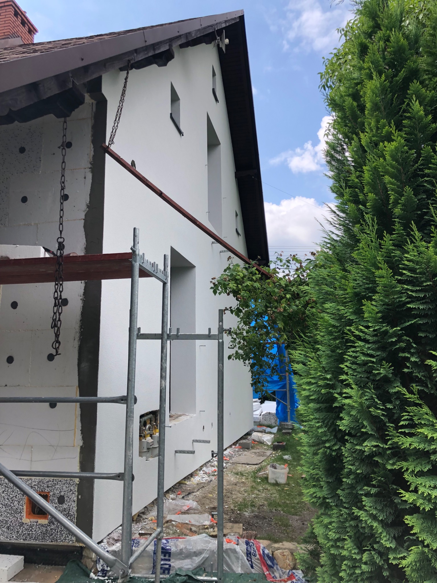 Dom w trakcie ocieplania: fragment elewacji z widoczną warstwą izolacji, rusztowanie z łańcuchami, fragment świeżo otynkowanej ściany oraz iglak w tle.