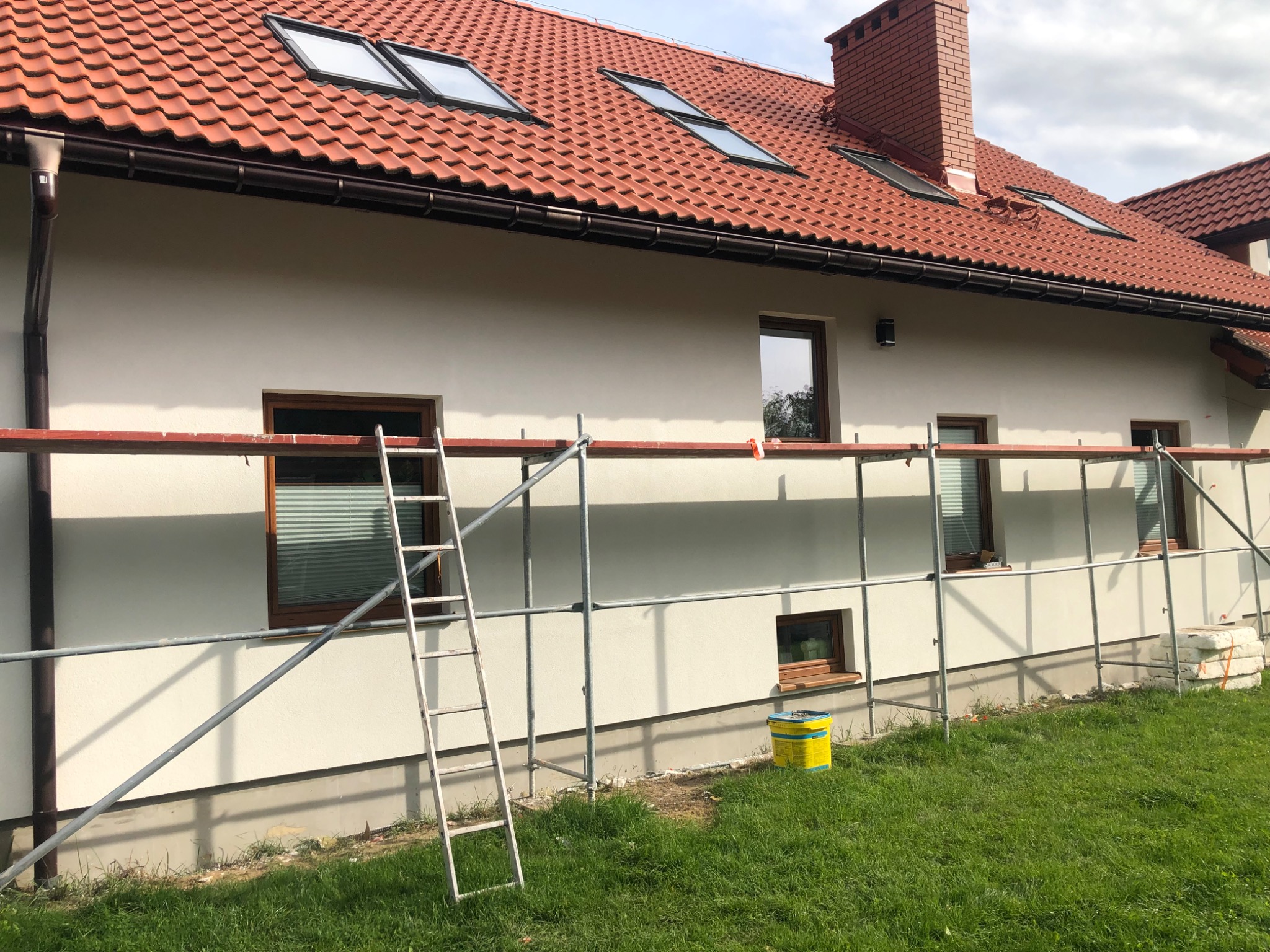 Elewacja domu jednorodzinnego w trakcie prac wykończeniowych, widoczne rusztowanie, drabina i wiadro z farbą przy ścianie.