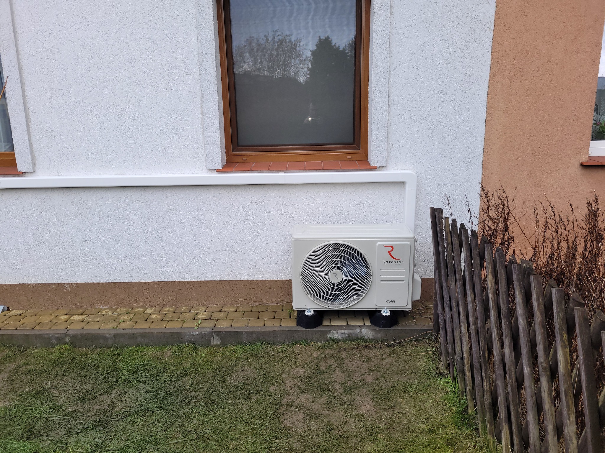 Biała jednostka zewnętrzna klimatyzacji Rotenso Ukura zamontowana na czarnych podkładkach, przy ścianie budynku z oknem w drewnianej ramie, widoczna drewniana palisada i trawa.