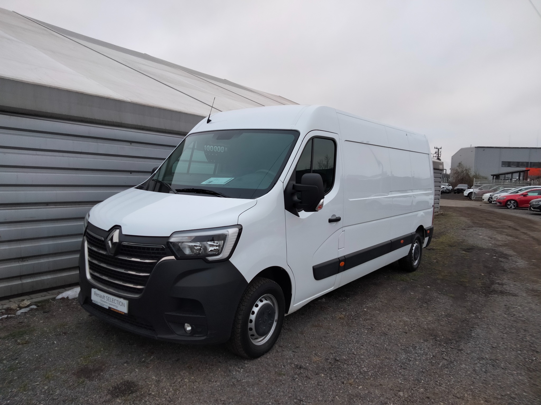 Biały Renault Master, nowy, stojący na placu z szarym żwirowym podłożem, widok z przodu i boku, z budynkiem w tle.