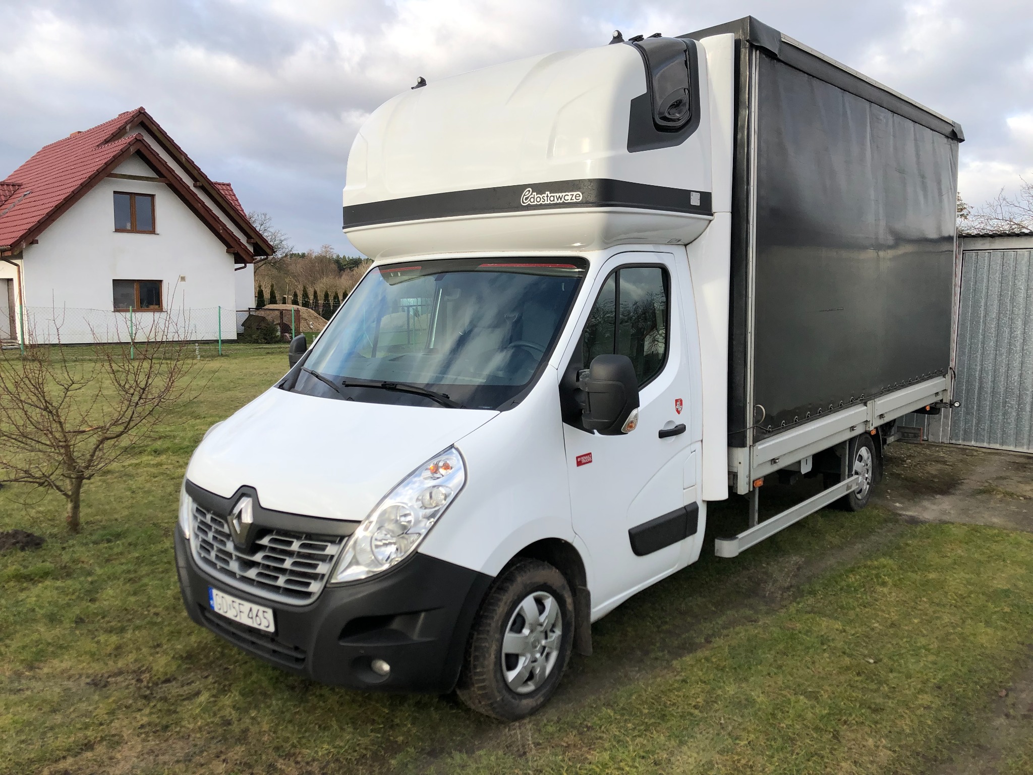 Biały samochód dostawczy Renault Master z plandeką, zaparkowany na trawniku przed domem z czerwoną dachówką, widoczny z przodu pod kątem.