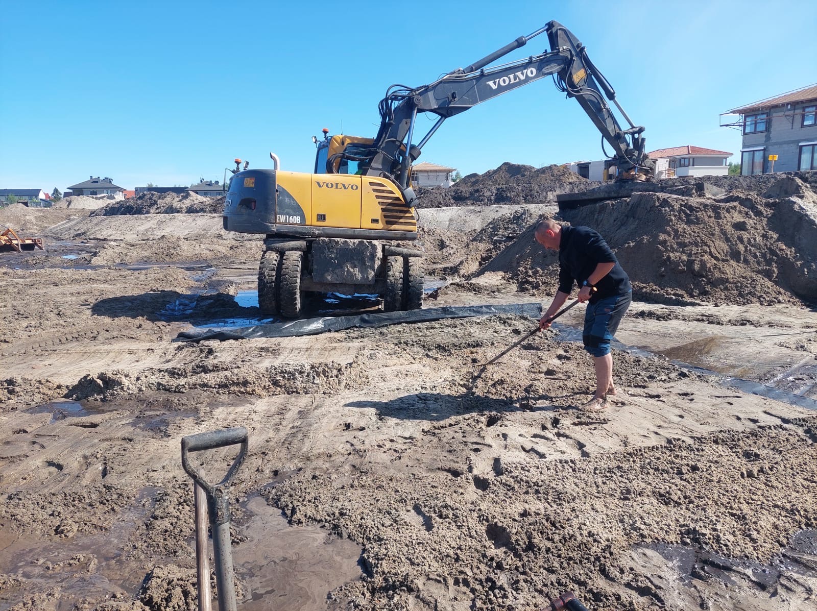 Żółta koparka Volvo EW160B na placu budowy, mężczyzna wyrównuje teren łopatą. W tle widać pryzmę piasku i budynki w trakcie budowy.
