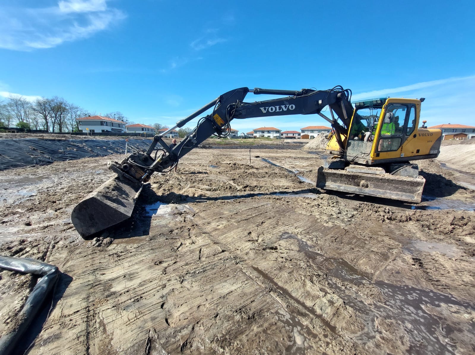 Żółta koparka Volvo na placu budowy z wykopanym terenem i widocznymi domami w tle, pod błękitnym niebem. Prace ziemne w toku.